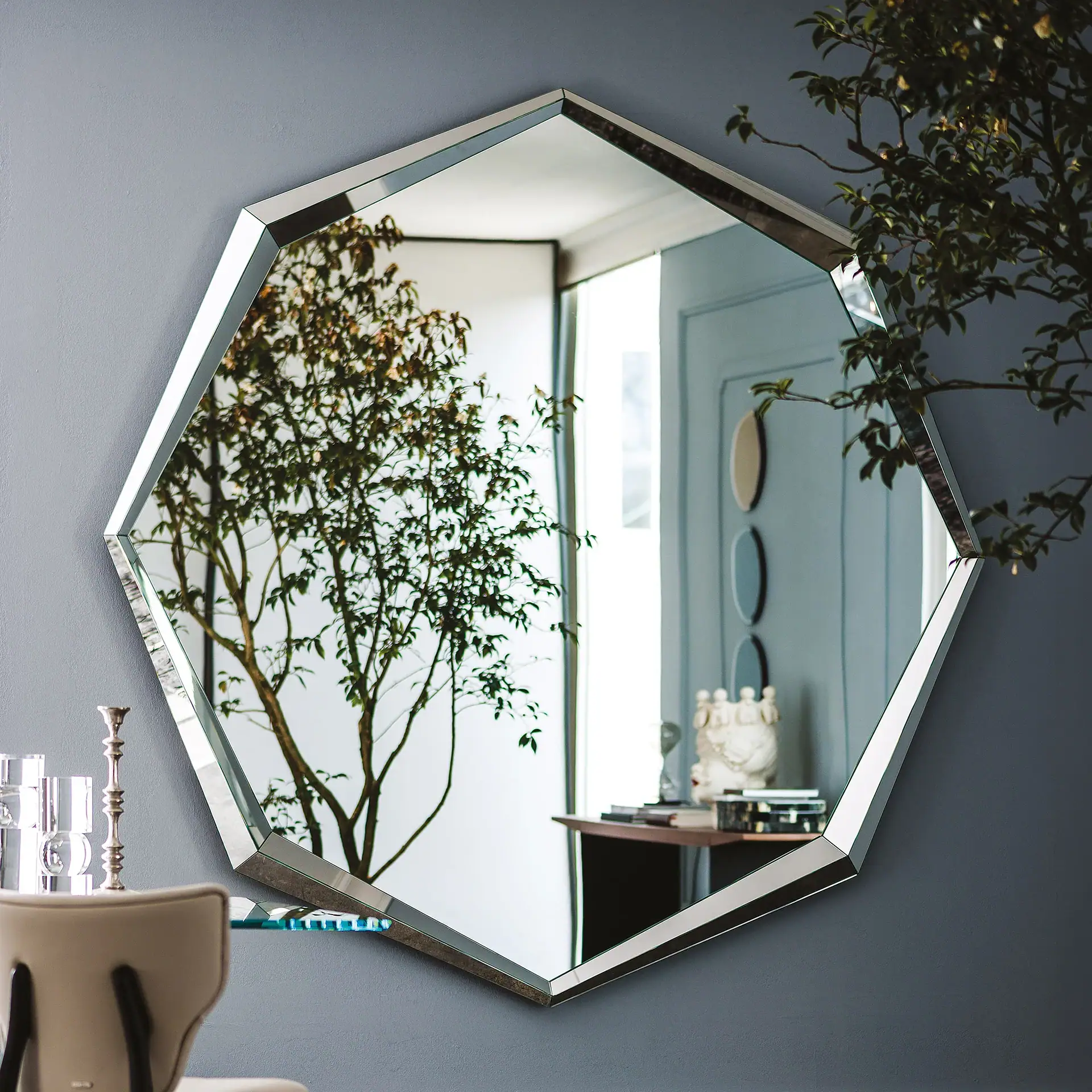 An octagonal mirror with a beveled edge hangs on a gray wall, reflecting a leafy green plant, a light blue paneled wall with oval decor, and a small table with a white sculpture.