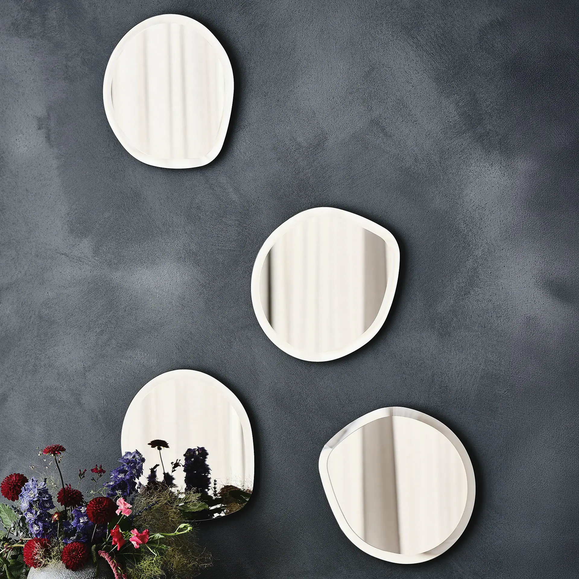 Four irregularly shaped mirrors on a dark gray textured wall, reflecting light. A vibrant floral arrangement with red, purple, and pink flowers is in the bottom left.