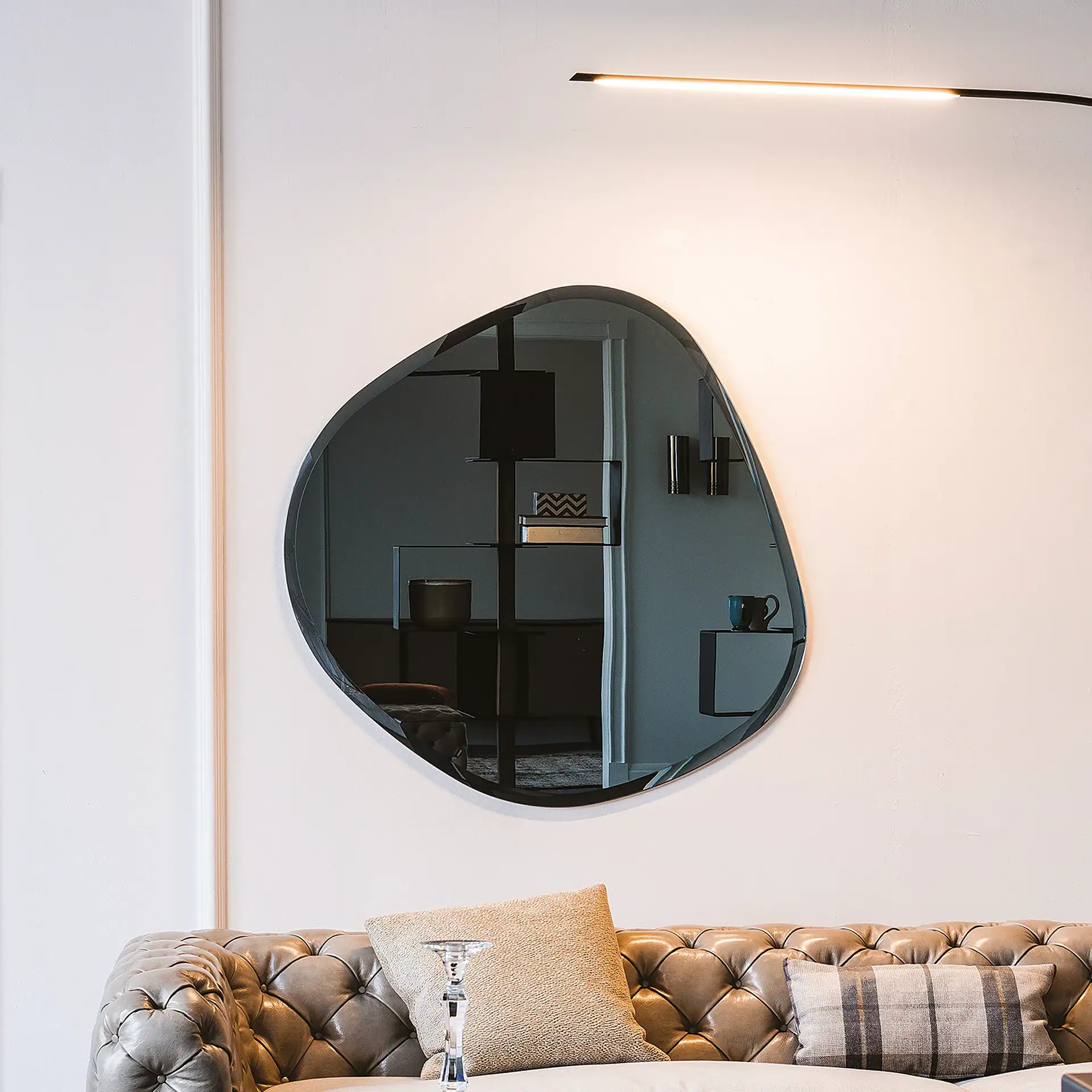 A large, irregularly shaped dark mirror on a light wall above a tufted leather sofa. A linear LED light is above the mirror, which reflects a modern shelving unit.