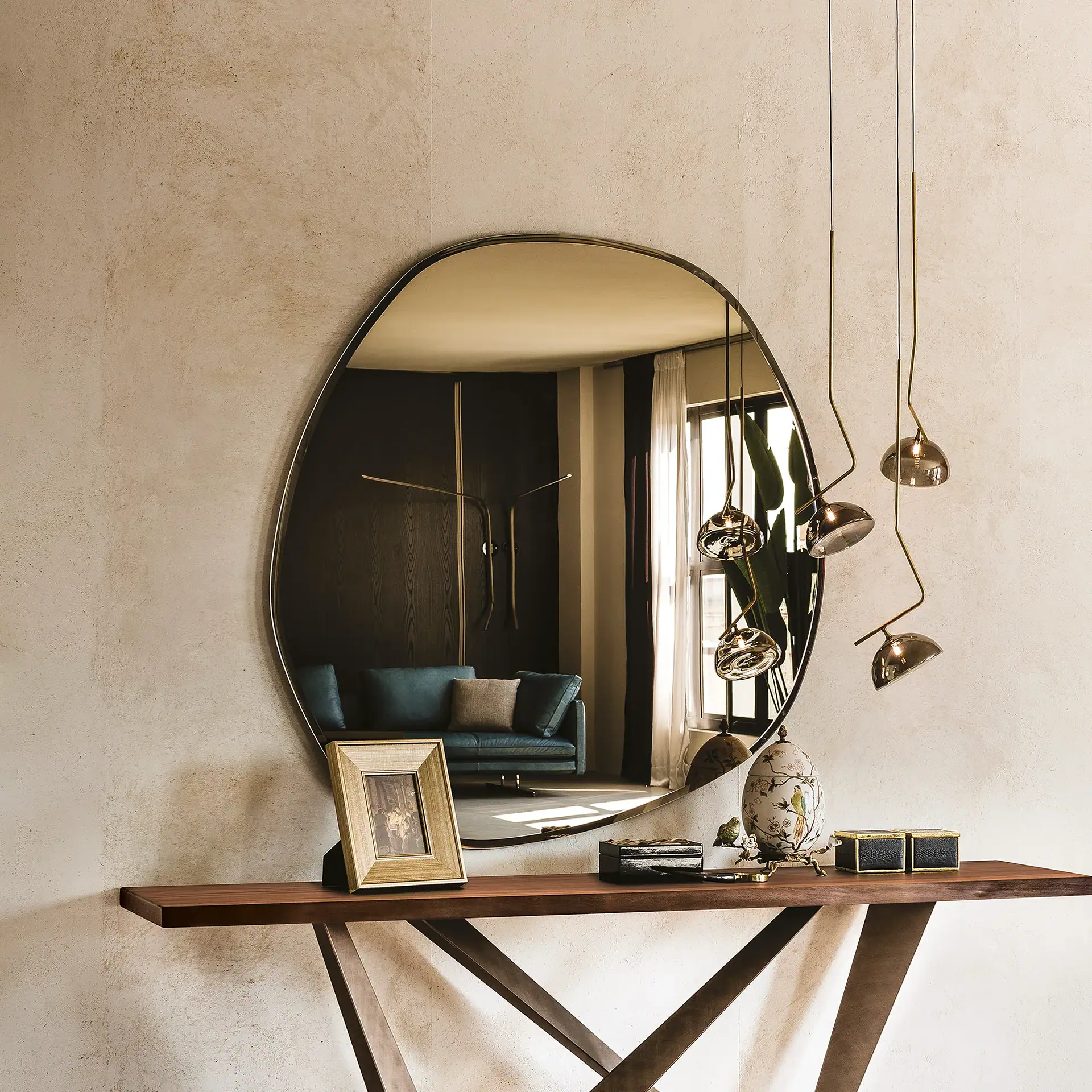 An oval mirror above a wooden console table with decorative items and modern pendant lights in an elegant interior.