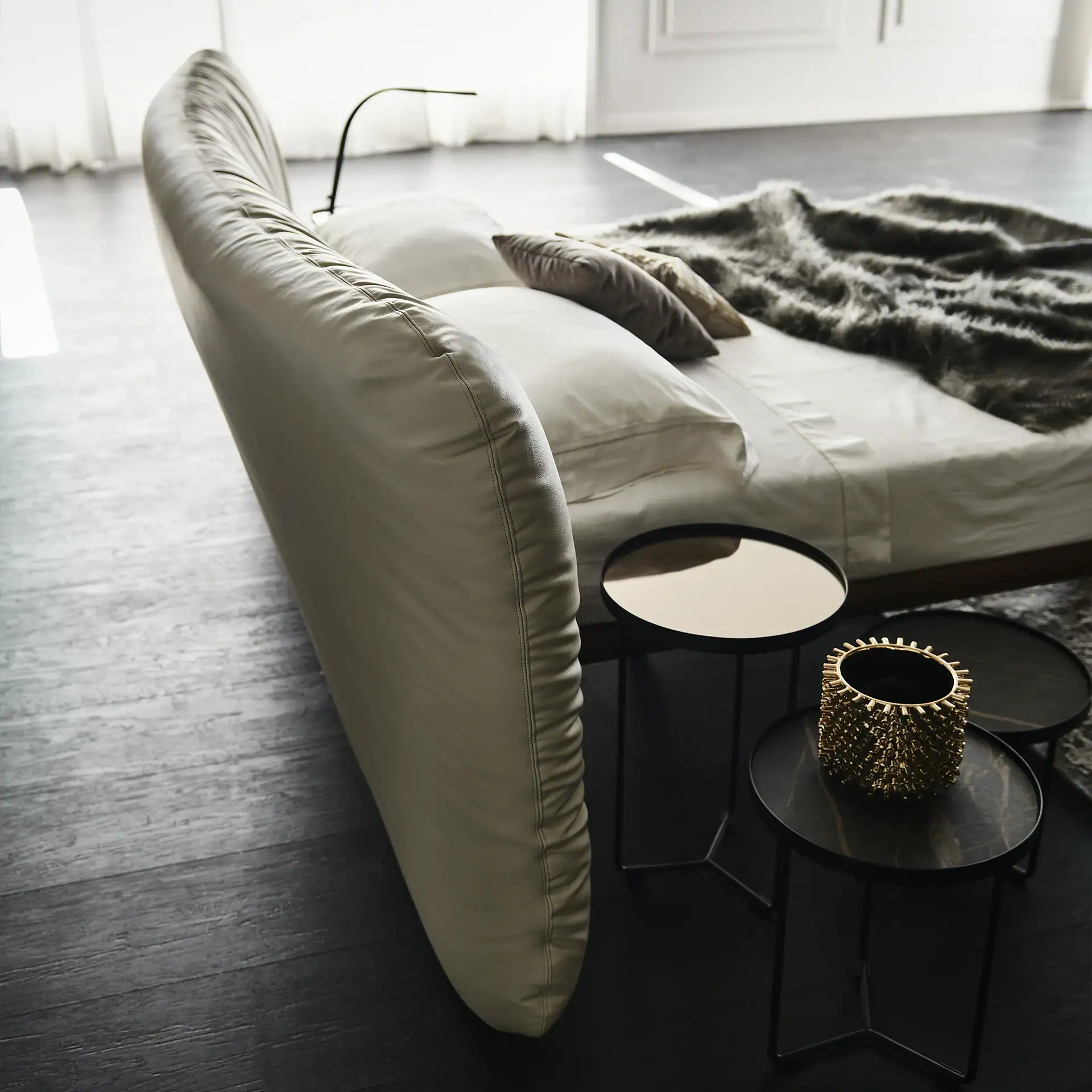 A modern bedroom with a large, curved beige leather bed, white bedding, dark faux fur throw, and two round side tables with a gold vase.