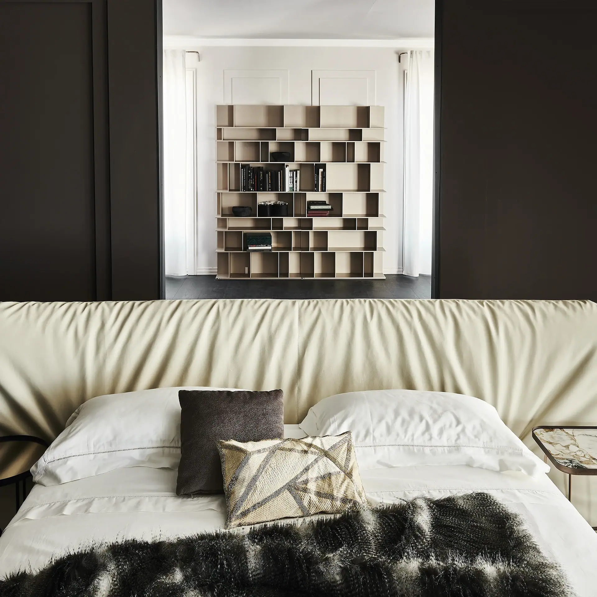 A modern bedroom with a cream upholstered headboard, white bedding, and a dark faux fur throw, looking into another room with a large, geometric light-colored bookcase.
