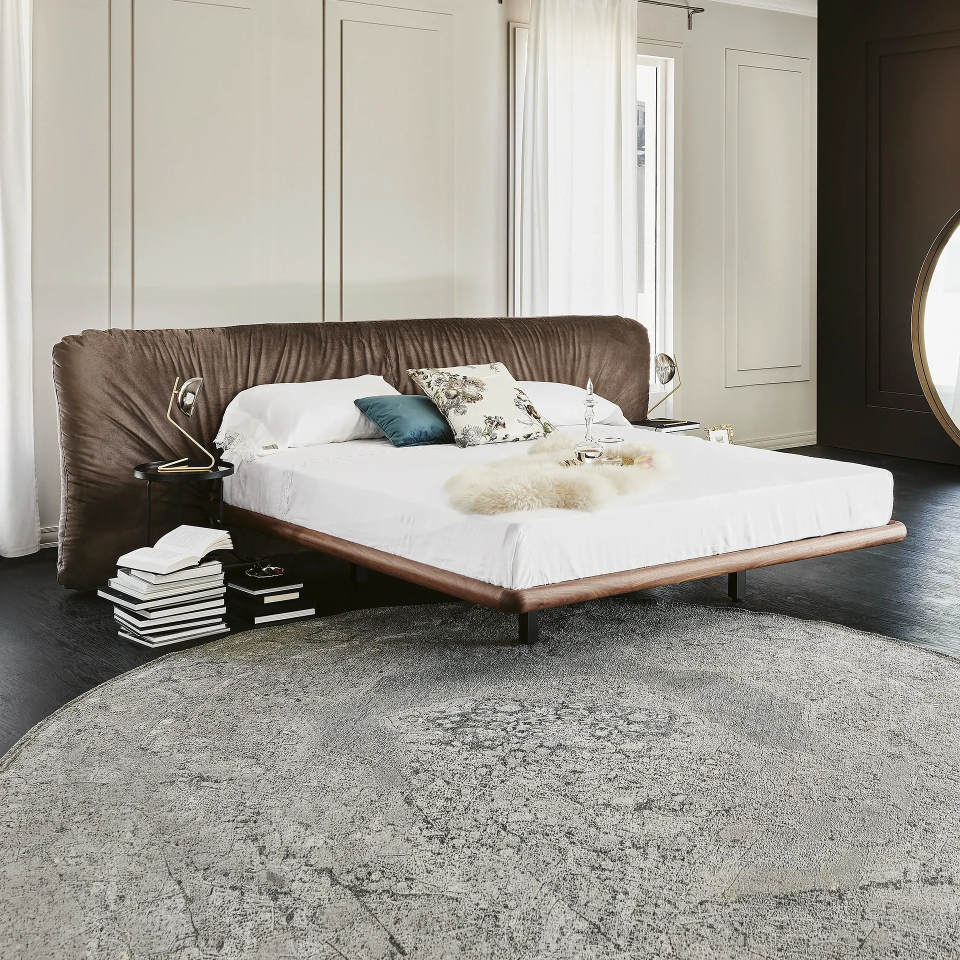A modern bedroom with a large bed featuring a dark brown plush headboard, white bedding, a round grey rug, and a gold bedside lamp.