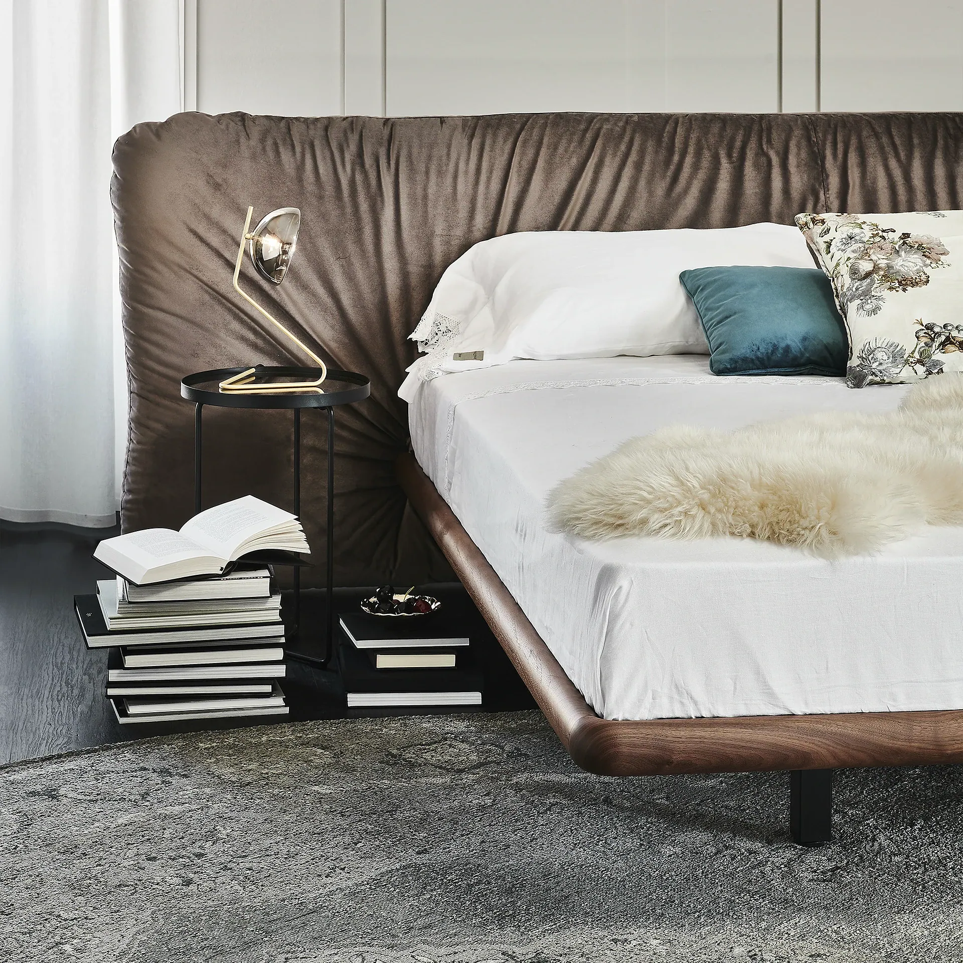 A modern bed with a dark brown velvet headboard, white sheets, and decorative pillows. A gold lamp sits on a black side table next to stacks of books on a dark floor with a gray rug.
