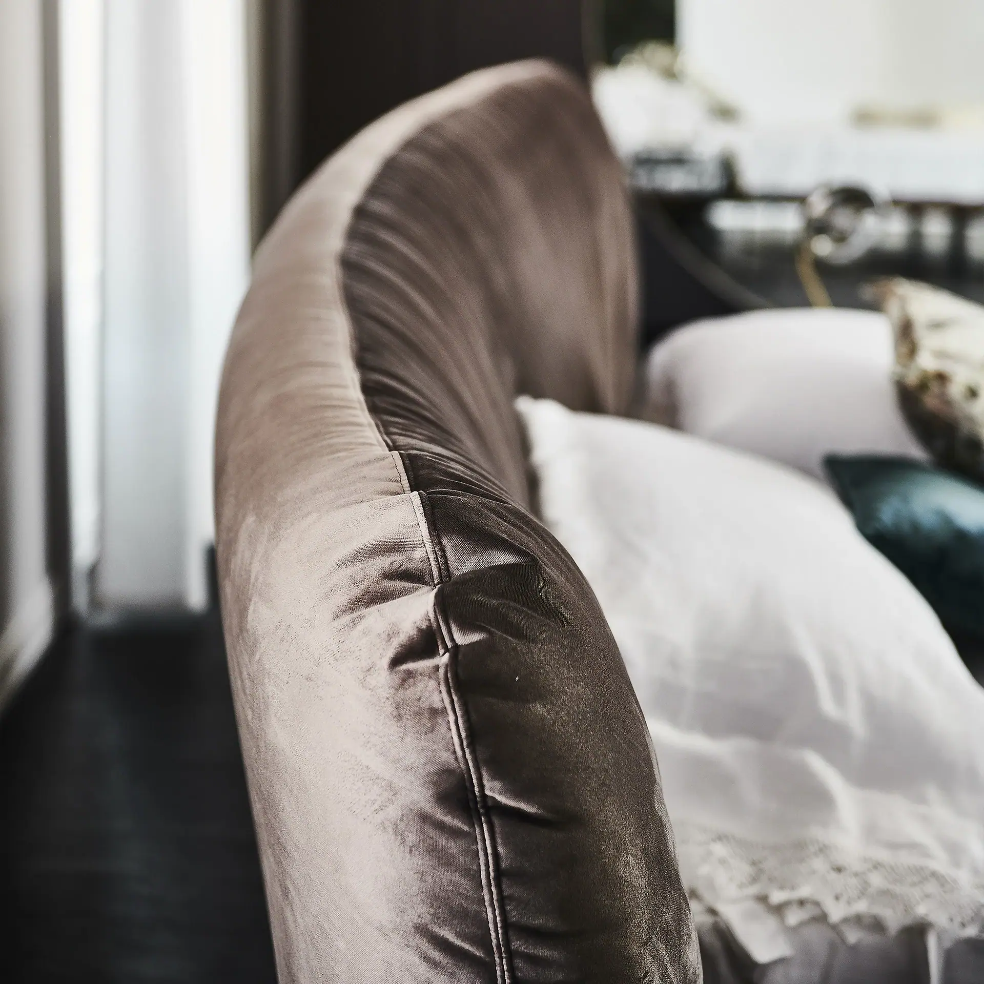 Close-up of a curved, brown velvet sofa armrest or backrest, with white and teal pillows in the soft-focus background.