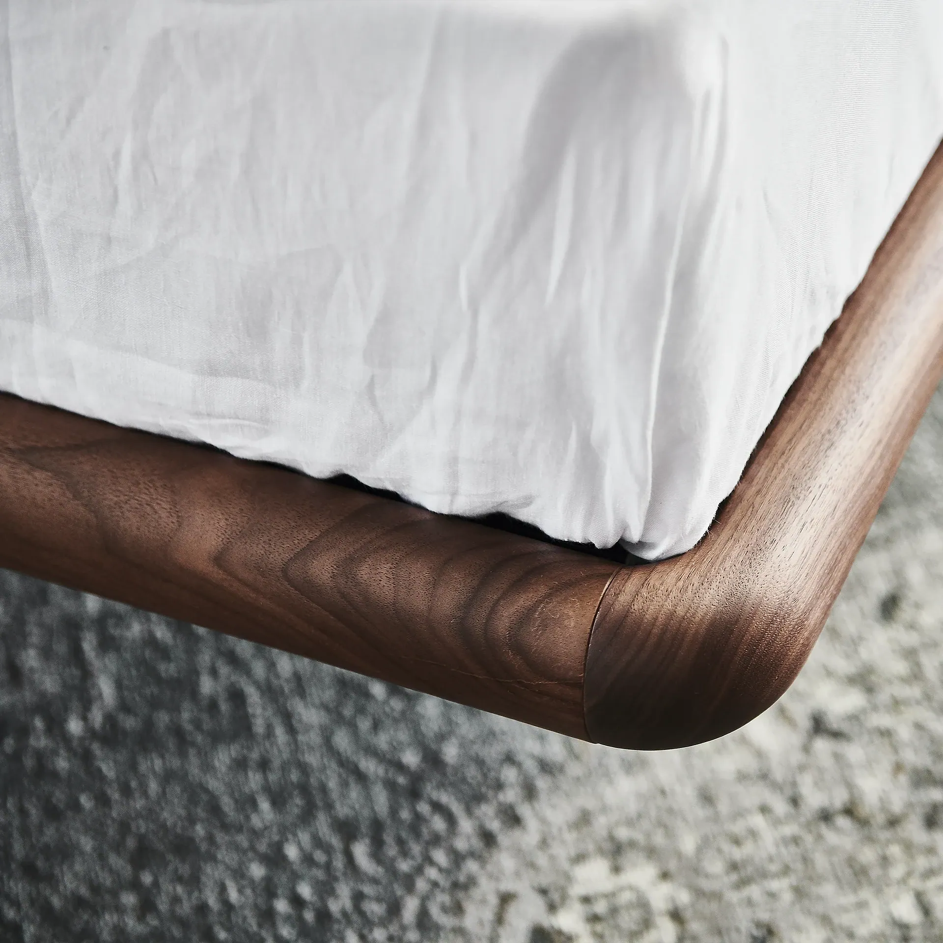 Close-up of a bed corner with a white wrinkled sheet and a dark brown rounded wooden frame over a blurred gray rug.