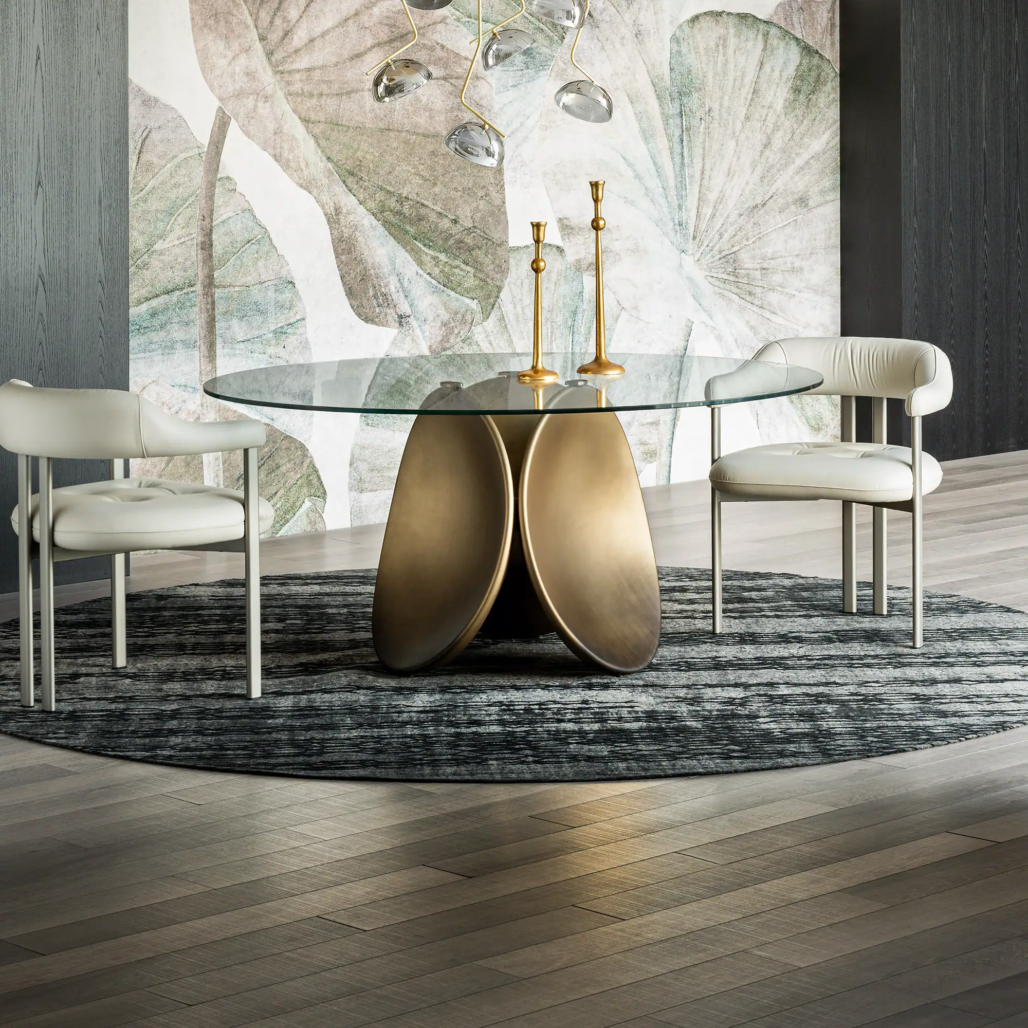 A modern dining room with a round glass table, bronze base, two light-colored chairs, a dark patterned rug, and a botanical wallpaper.