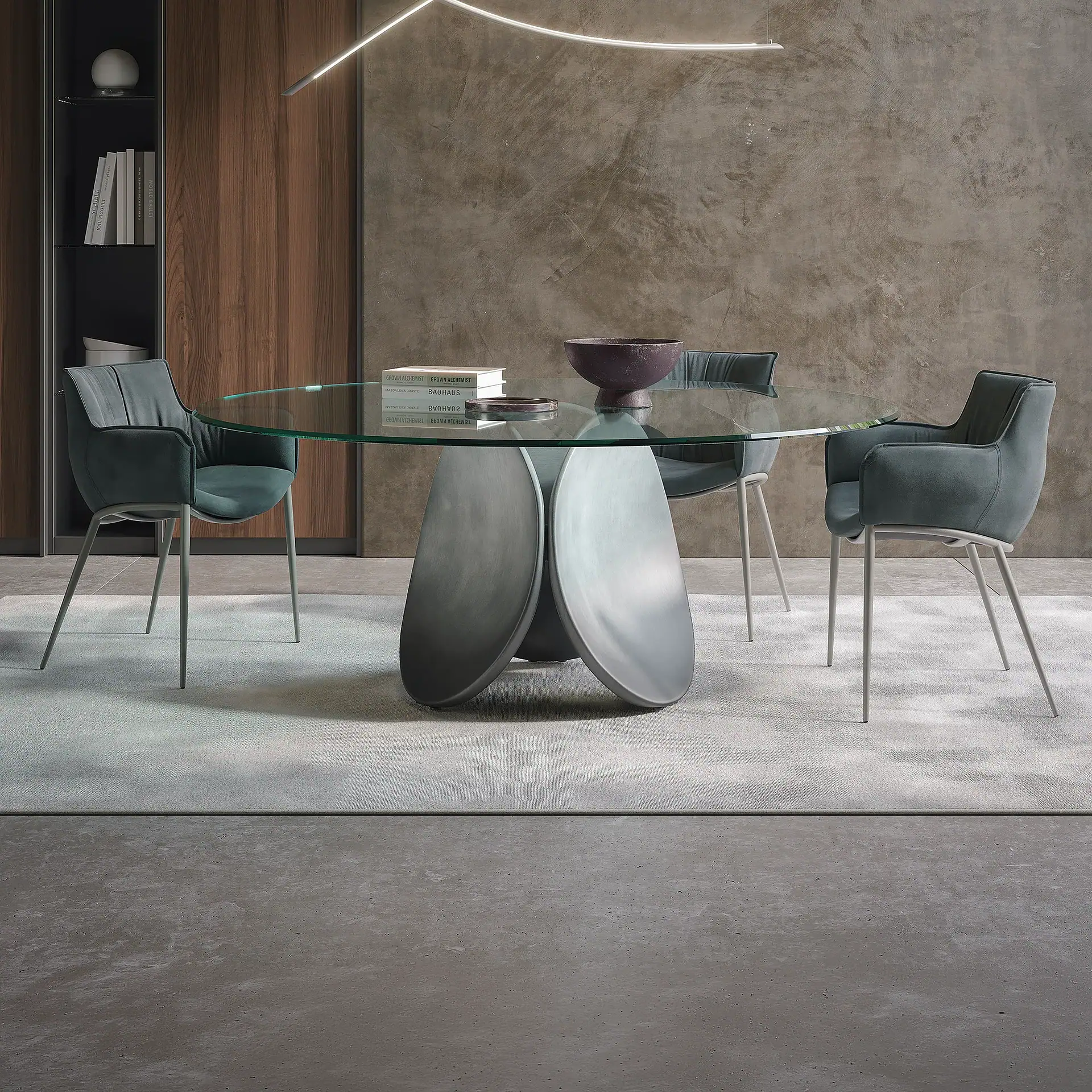 A modern dining room with a round glass table, a sculptural grey base, and three dark teal upholstered chairs on a light grey rug. A textured grey wall and a dark wood shelving unit are in the background.