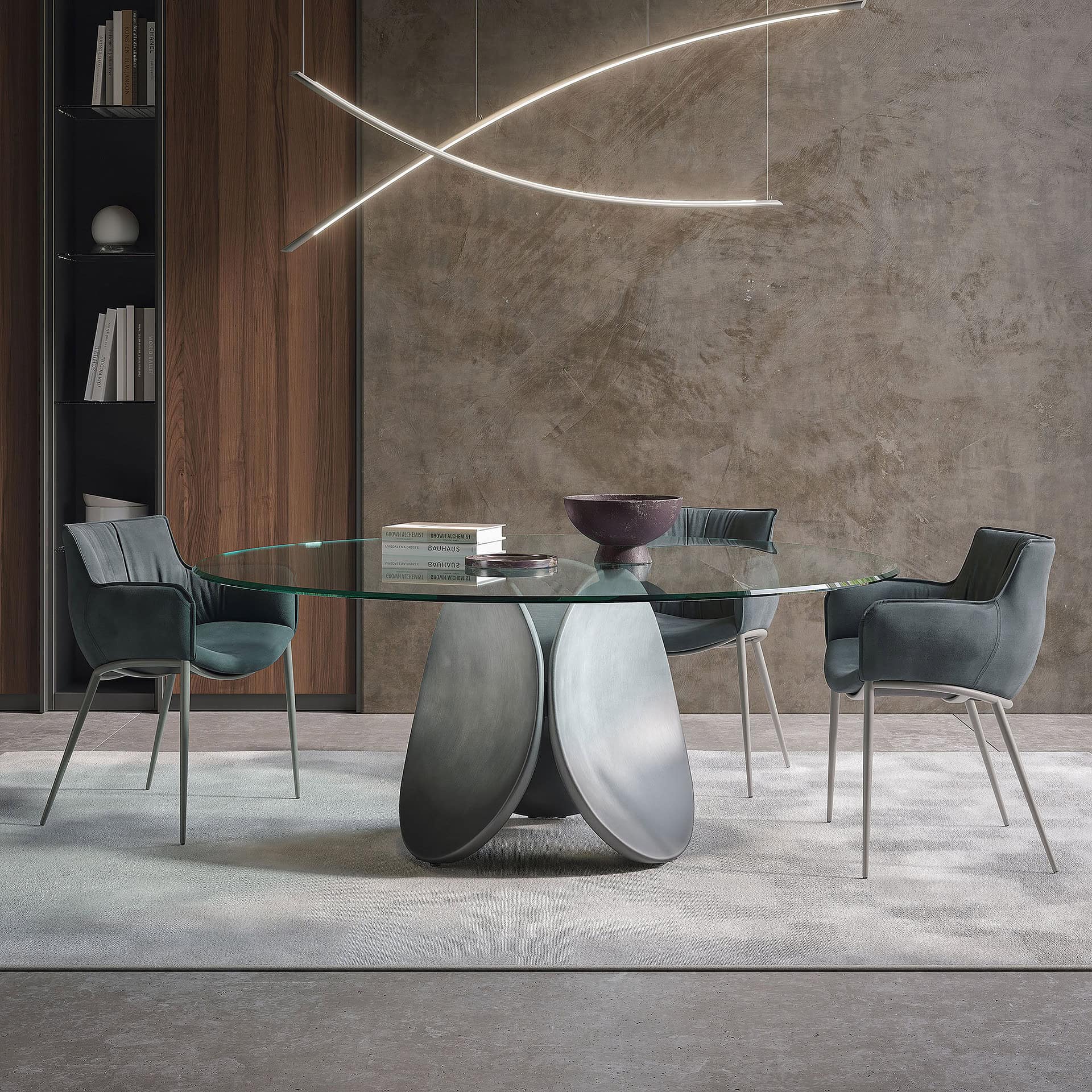 A modern dining room with a round glass table, sculptural metal base, four dark teal chairs, and a curvilinear pendant light. A textured concrete wall and a wooden cabinet with books are in the background.