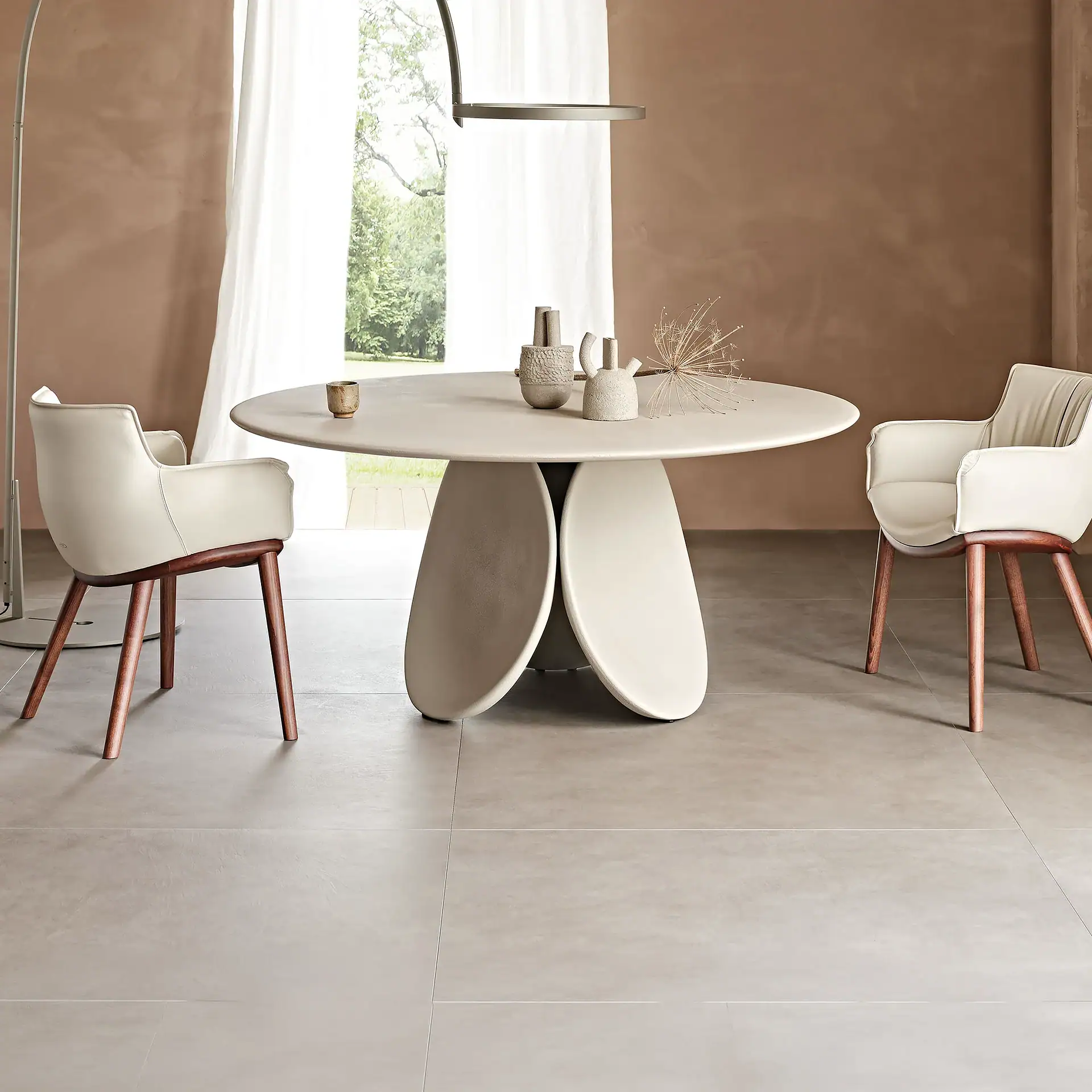 A modern dining room with a light beige oval table, two cream chairs with wooden legs, and minimalist decor. A large window with sheer curtains overlooks green trees.