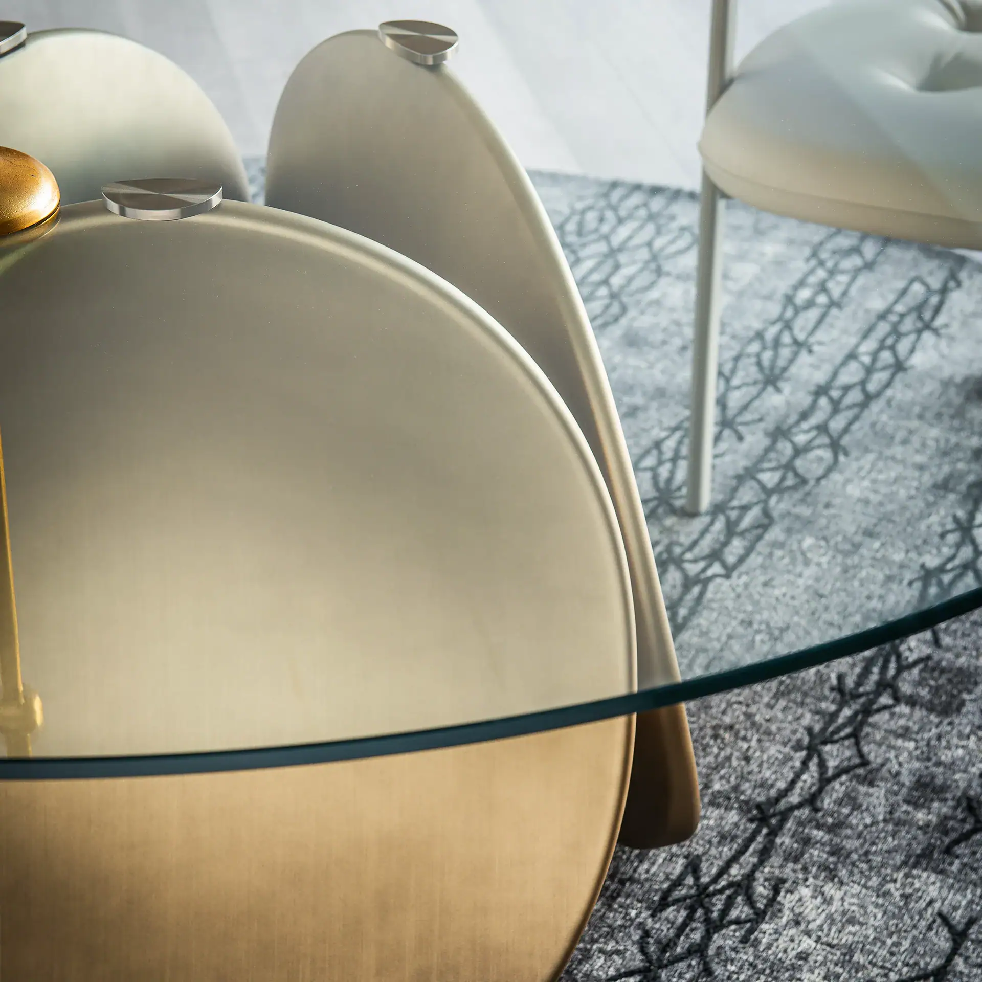 Close-up of a modern coffee table with a clear glass top and a brushed gold/brass curved base, on a grey patterned rug.