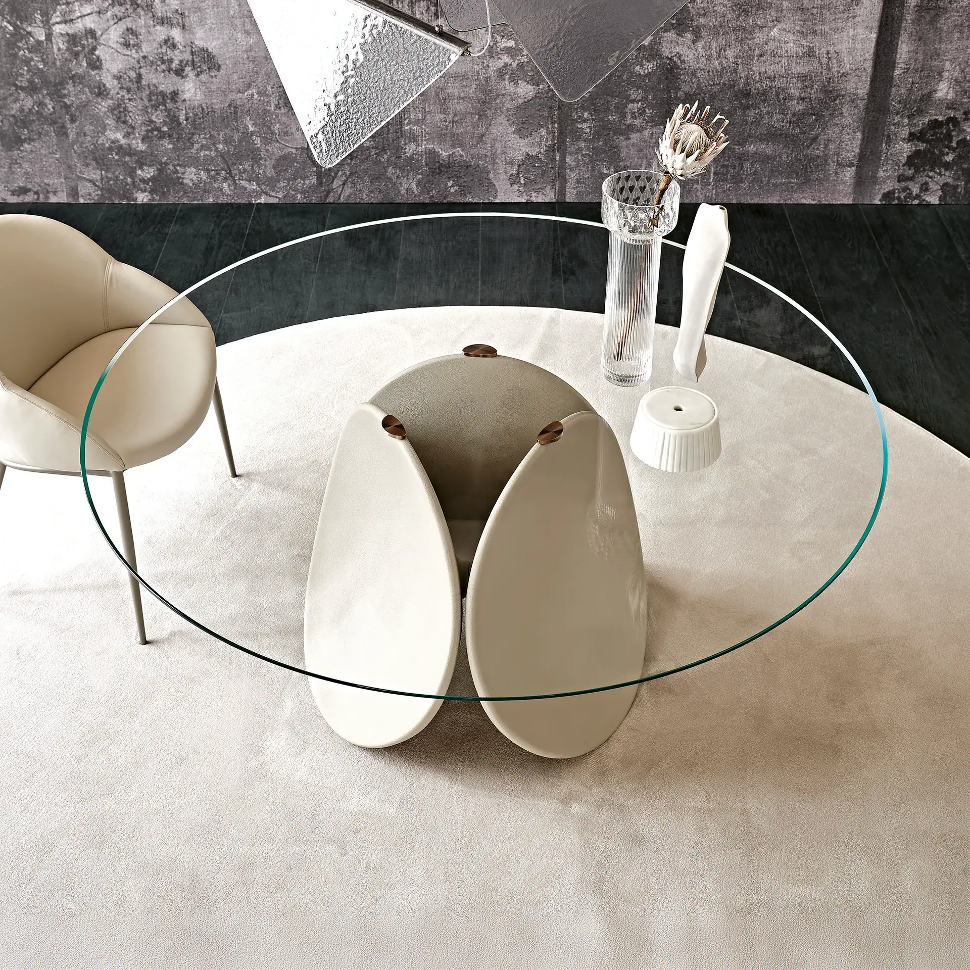 A modern living room scene with an oval glass coffee table, a cream chair, and a light beige rug on a dark floor. The table has a unique base made of three tear-drop shaped elements and is adorned with a dried protea flower in a vase, and white decorative objects.