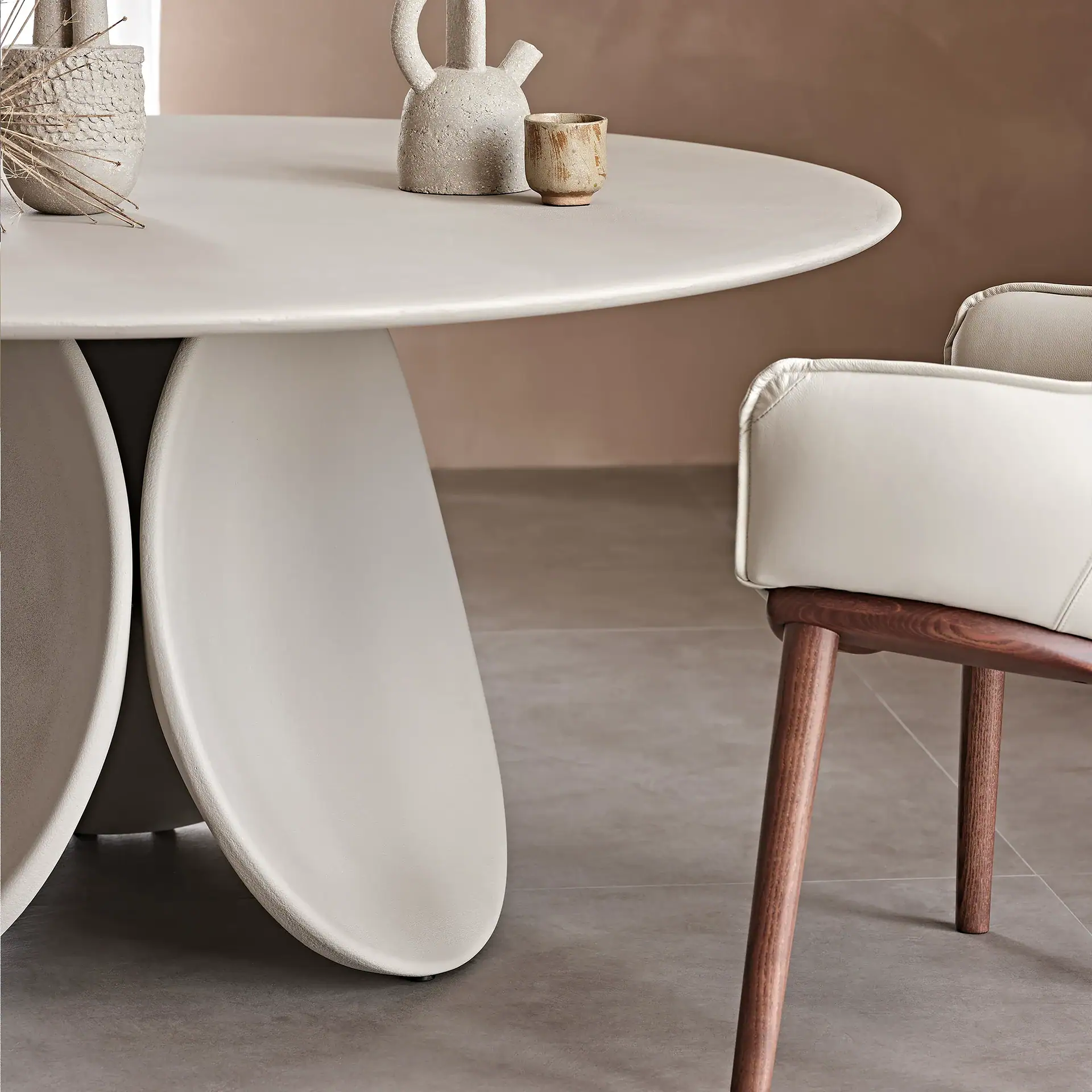 A round, light gray dining table with a unique panel base, adorned with ceramic decor and dried plants. A cream leather chair with wooden legs is visible on the right, all set against a muted brown wall and gray tiled floor.
