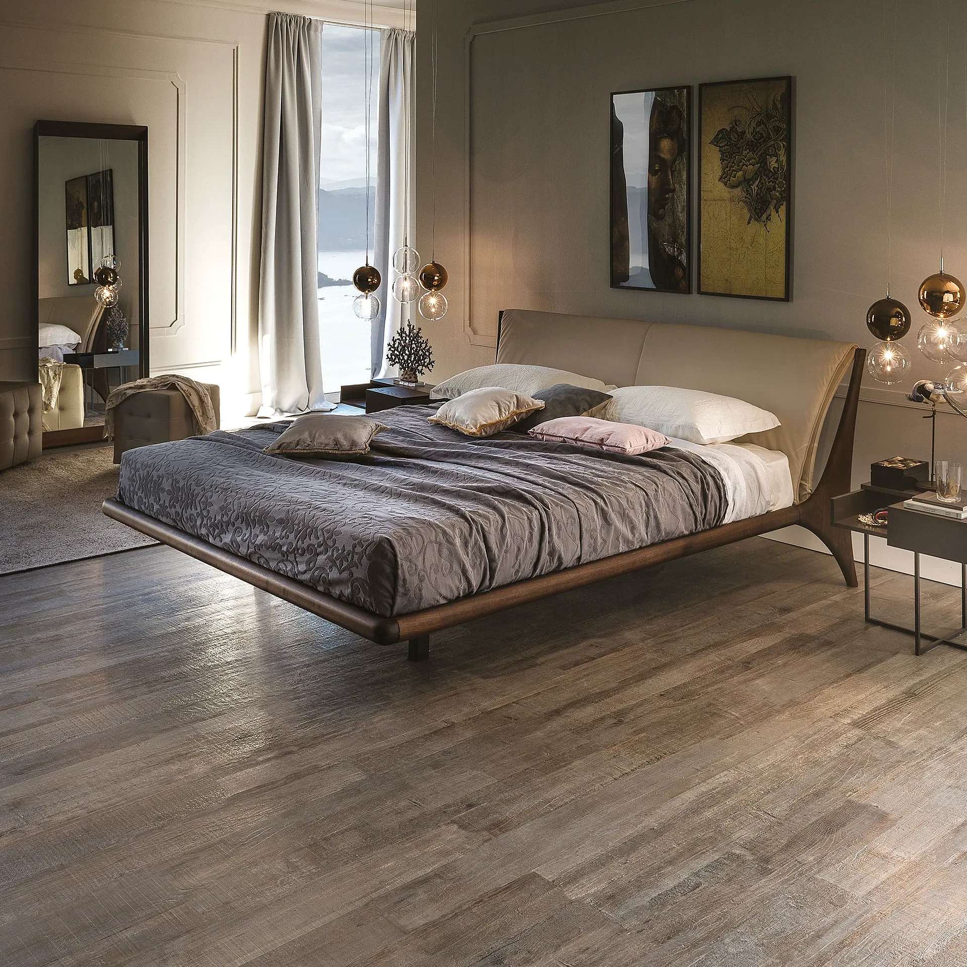 A modern bedroom with a floating bed, beige headboard, gray duvet, and multiple pillows. A large window overlooks a lake and mountains. Two framed artworks hang above the bed, and pendant lights illuminate the space. Wood-look flooring completes the room.