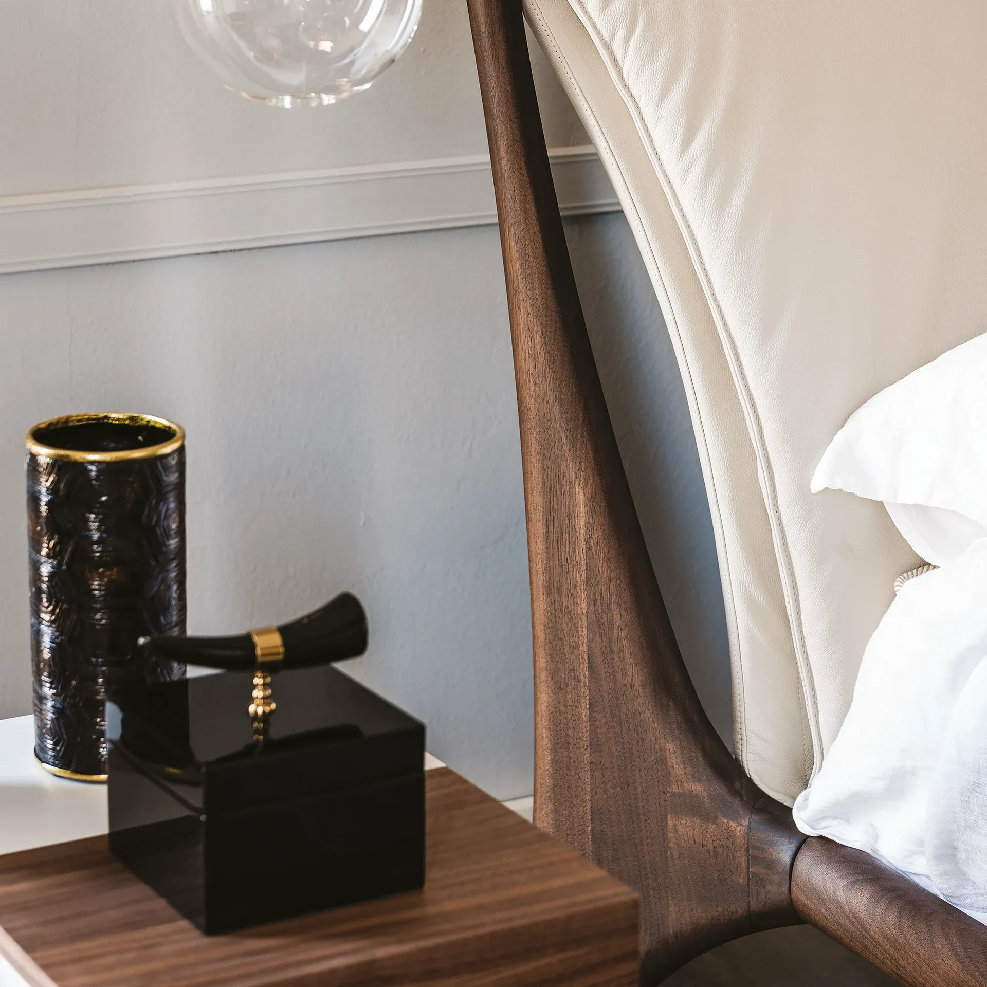 Close-up of a modern bedside table with a dark textured vase and a black decorative box, next to a bed with a curved wooden and cream upholstered headboard.