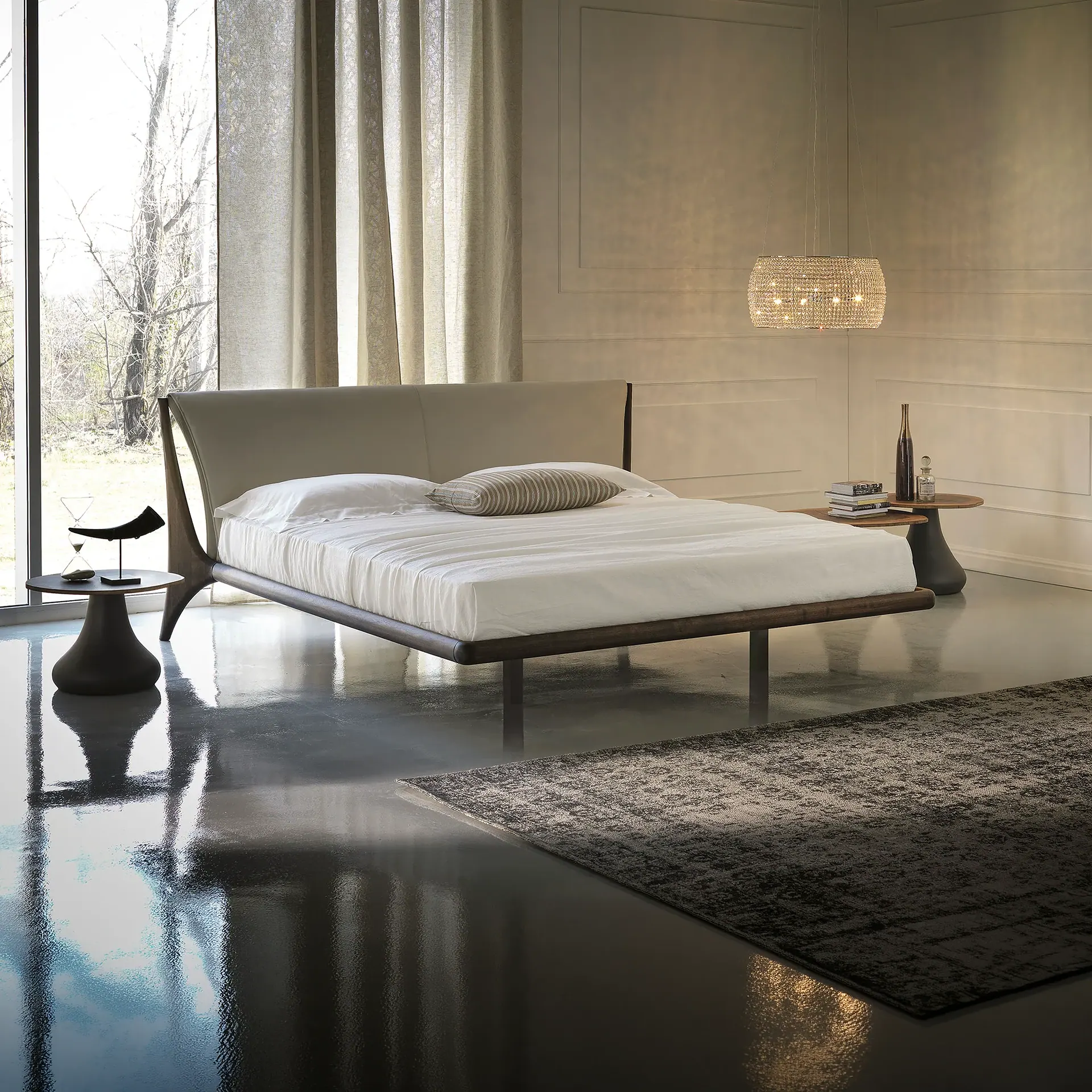 A modern bedroom with a low-profile bed, light upholstered headboard, large window with sheer curtains, reflective dark floor, and a sparkling crystal chandelier.