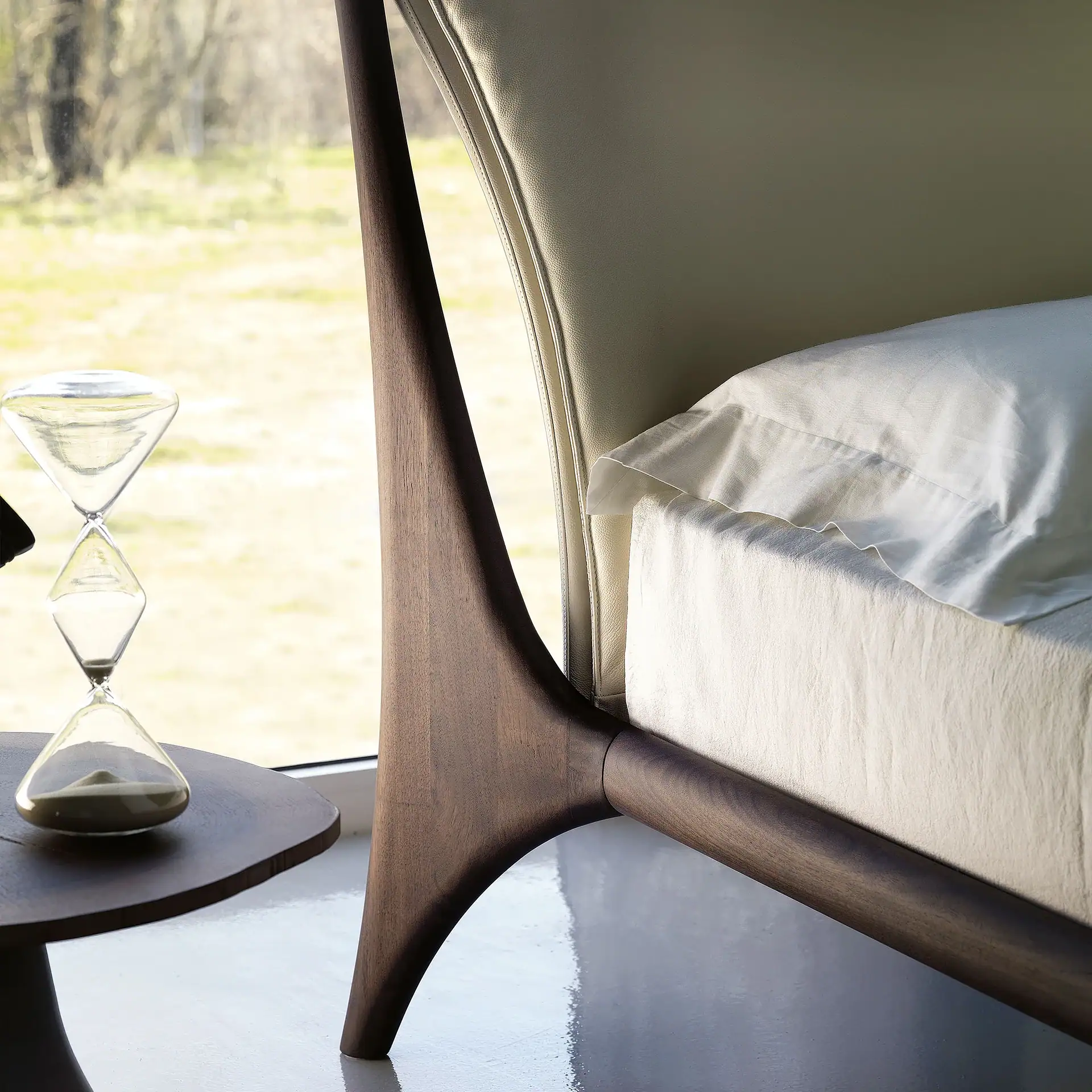 A modern bed with a dark wood frame and light leather headboard, white linen, and an hourglass on a wooden side table by a window with an outdoor view.