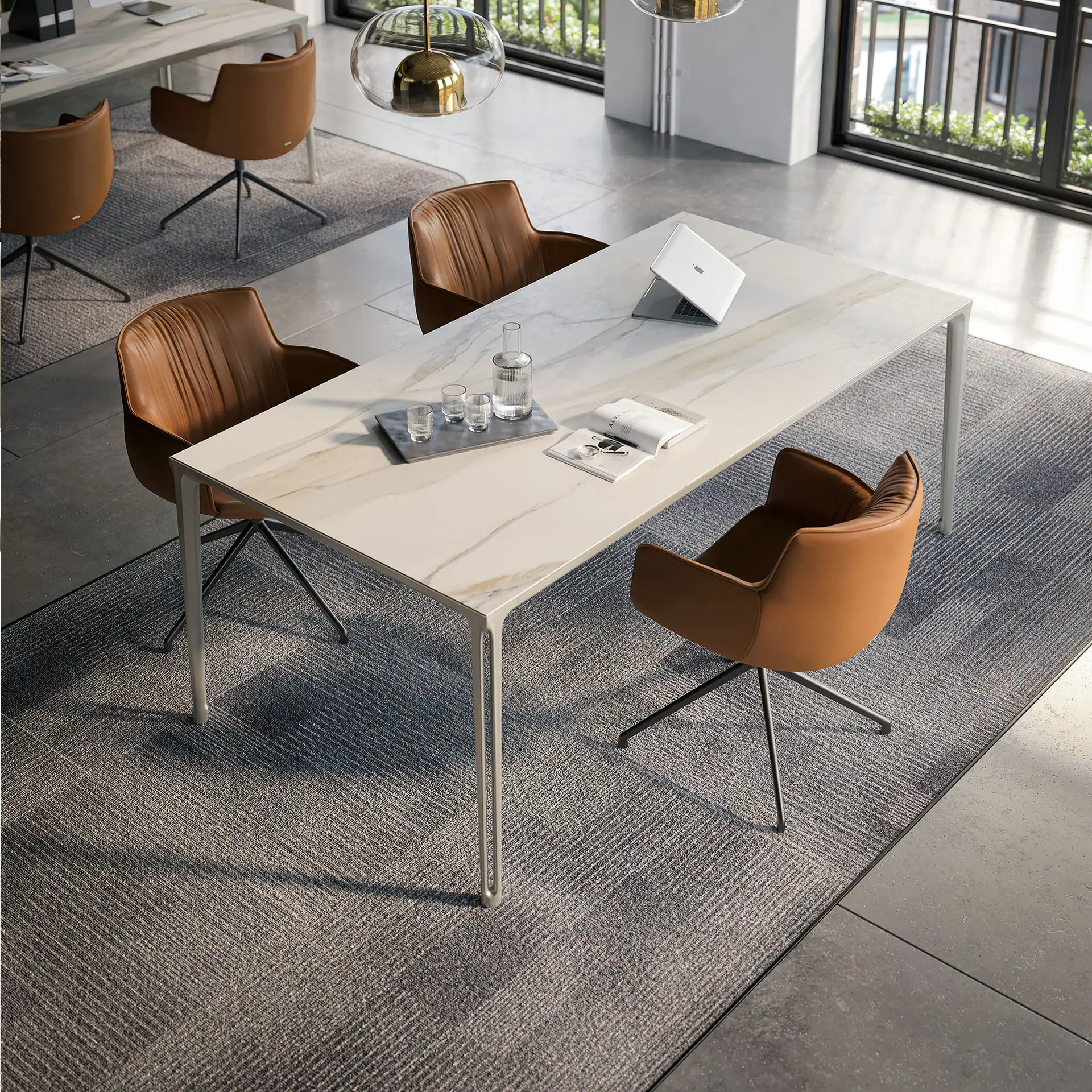 A modern office table with a marble top and brown leather chairs on a grey rug. A laptop, water carafe, and book are on the table. A large window with greenery outside is in the background.