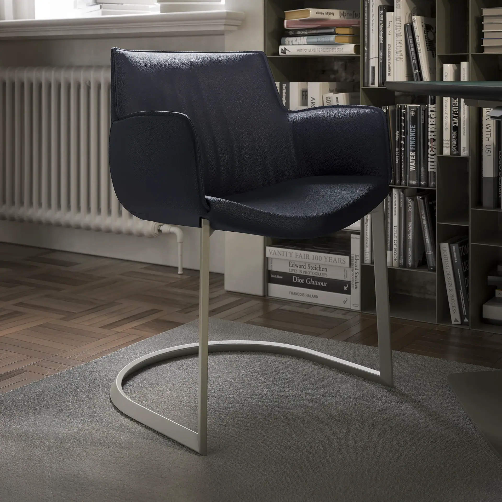 A dark blue leather armchair with a silver cantilever base on a grey rug, in a room with bookshelves filled with books and a parquet floor.