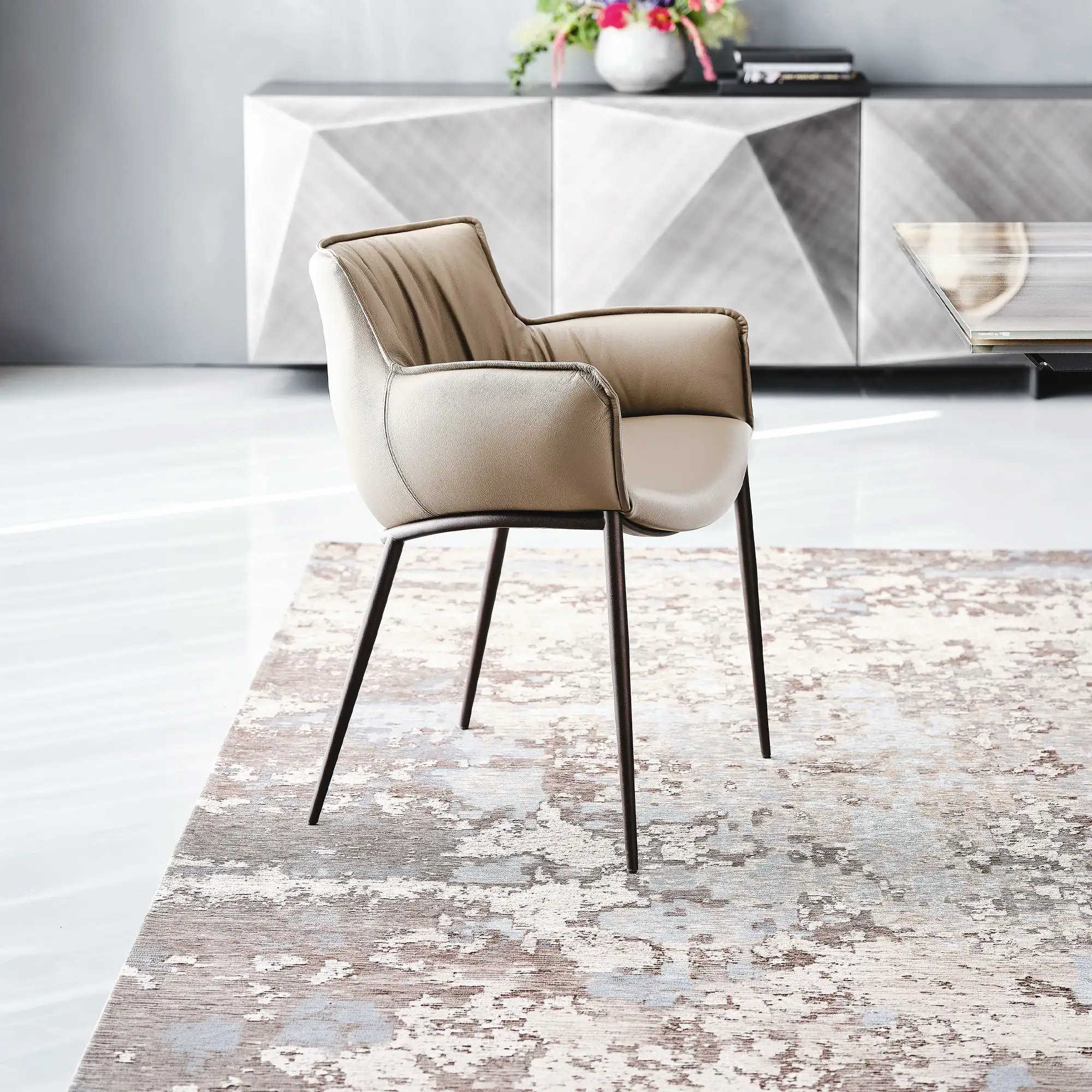 A modern beige leather armchair with dark metal legs sits on an abstract patterned rug in a room with a geometric grey credenza and floral arrangement.