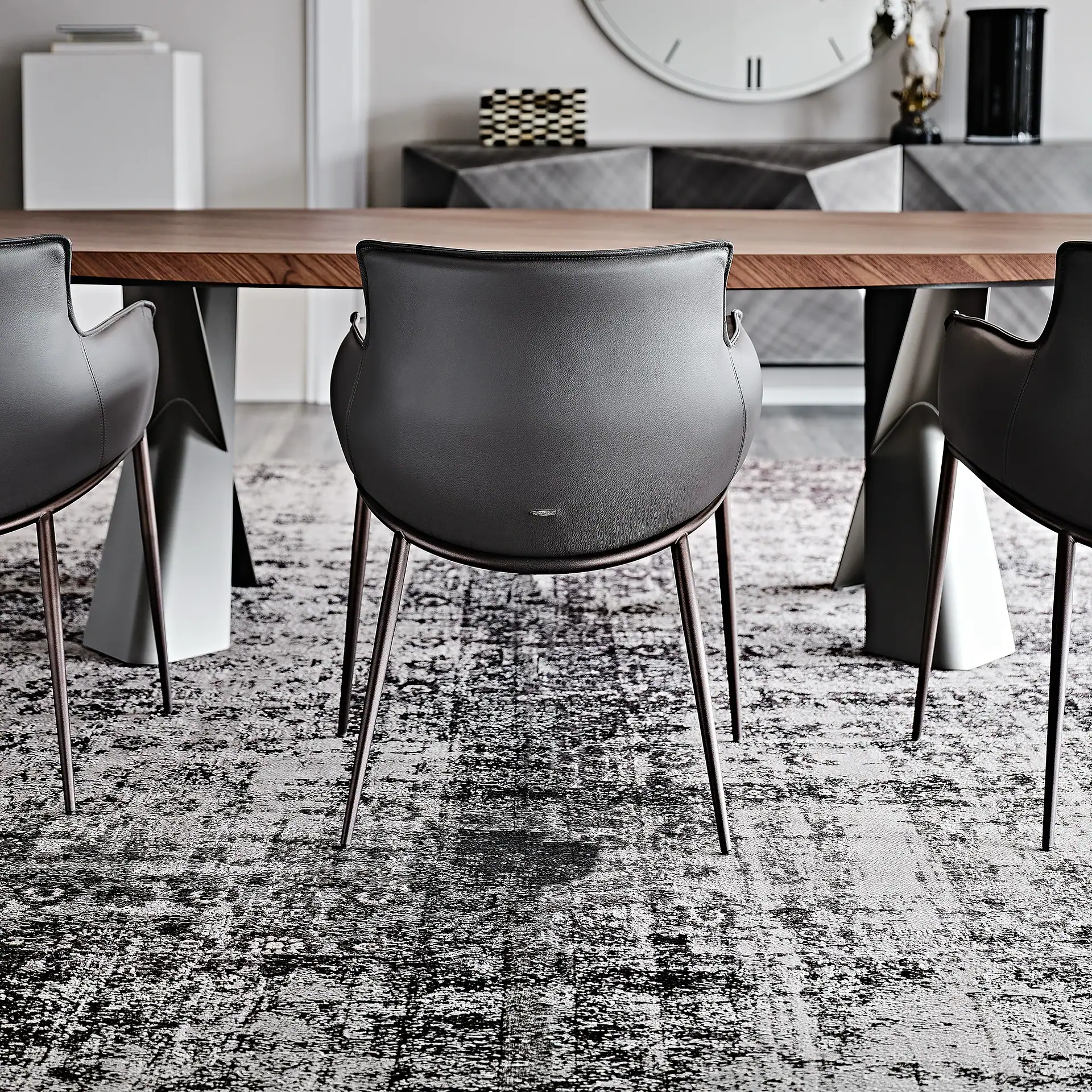 A modern dining room with a wooden table, dark gray leather chairs, and a patterned rug. A geometric sideboard and decorative items are in the background.