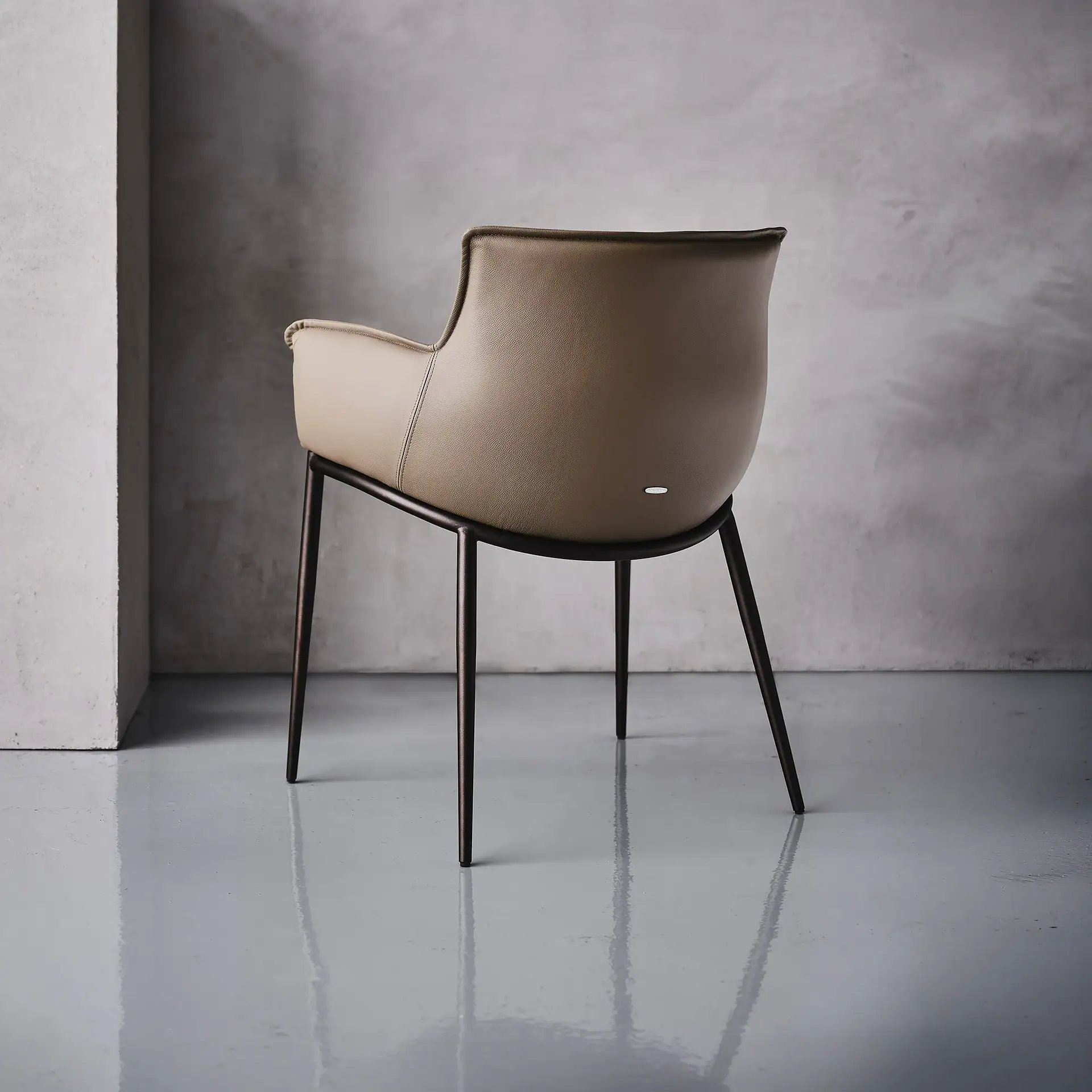 A modern dining chair with a light brown upholstered back and seat, and dark metal legs, against a grey wall and reflective grey floor.