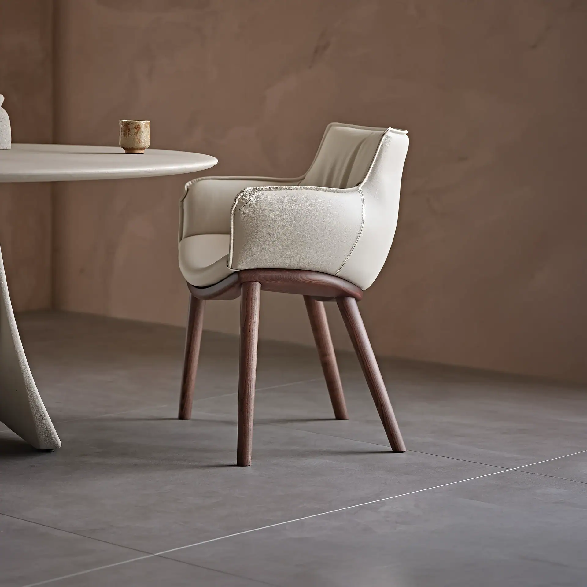 Cream upholstered dining chair with dark wooden legs next to a light gray round table with a ceramic cup. Dark gray tiled floor and warm brown wall.