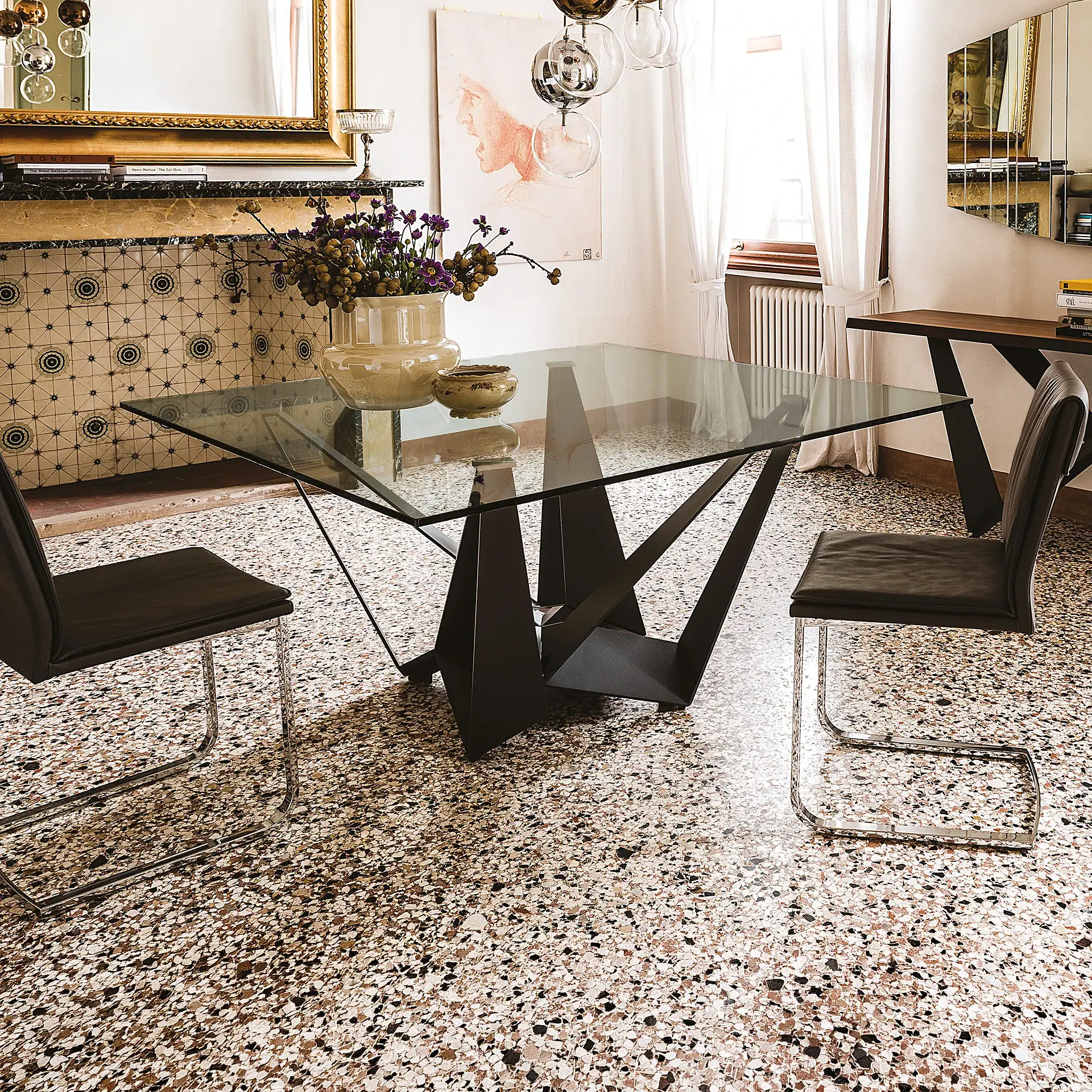 A modern dining room with a square glass table, two dark leather chairs, and a sculptural metal base on a terrazzo floor. A fireplace, gold mirror, and contemporary art adorn the walls.