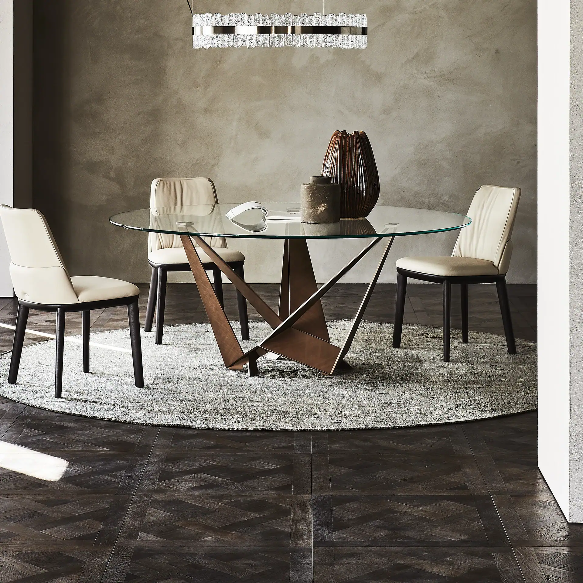 A modern dining room with a round glass table, four cream leather chairs, a gray rug, dark wood floor, and a crystal chandelier.
