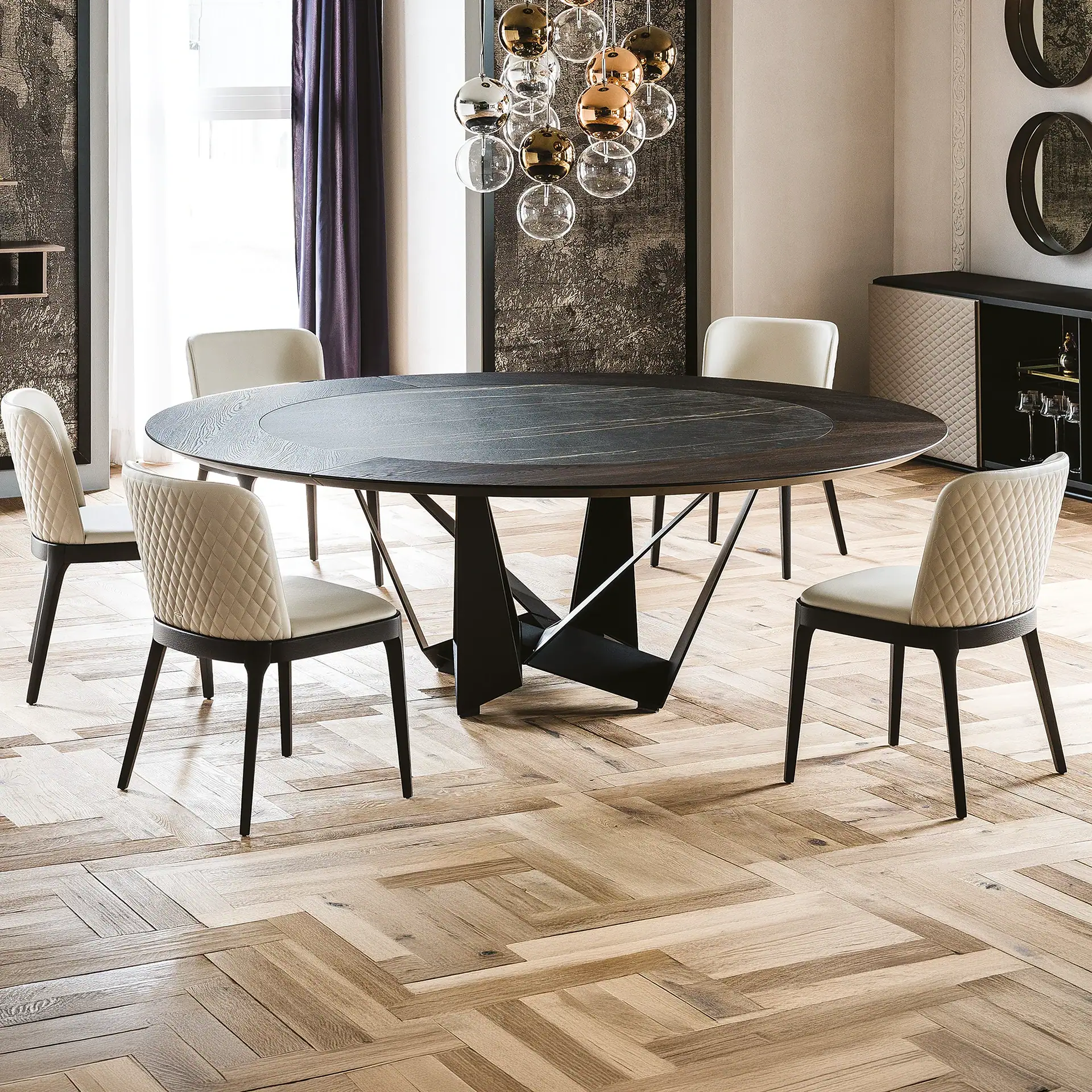 A modern dining room with a dark wood round table, six cream quilted chairs, a spherical chandelier, and herringbone wood flooring.