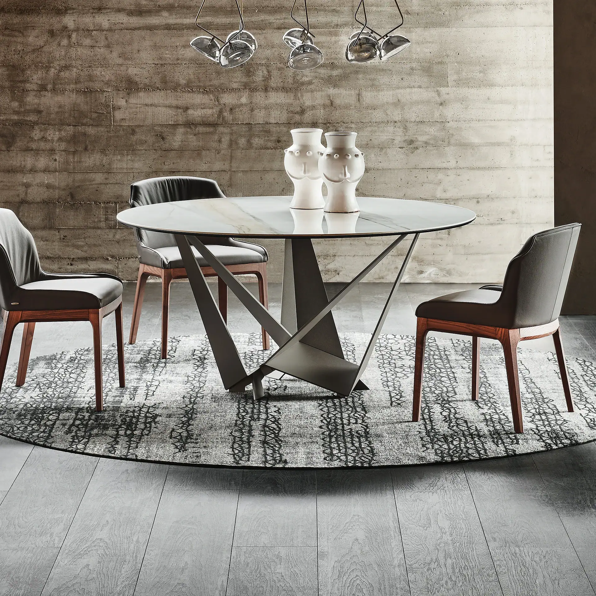 A modern dining room with a round marble table, three gray leather chairs, a round patterned rug, and a concrete wall.
