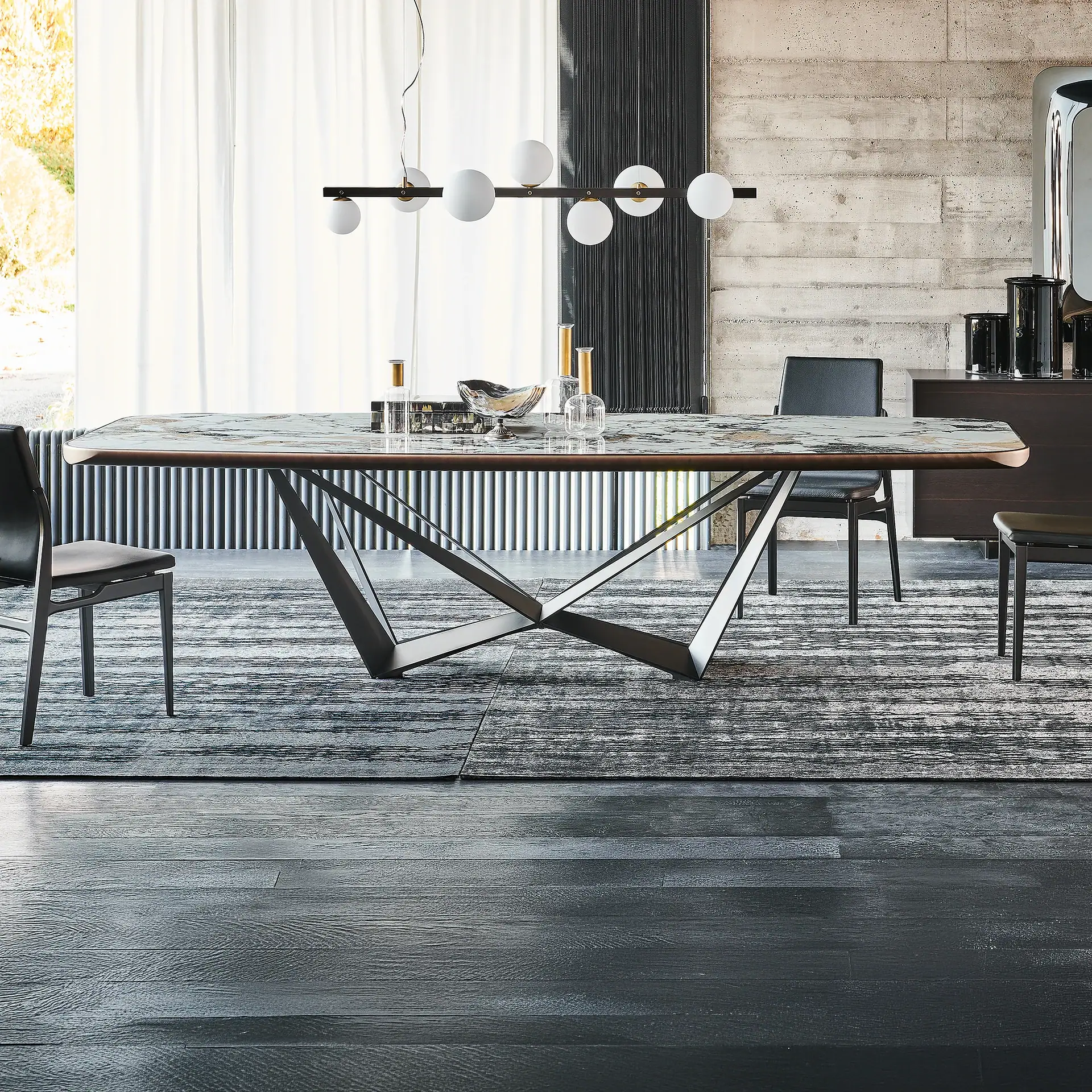 A modern dining room with a marble-top table, geometric metal base, and black leather chairs on a patterned rug. A linear chandelier hangs above, with sheer white curtains on the left and a concrete wall on the right.