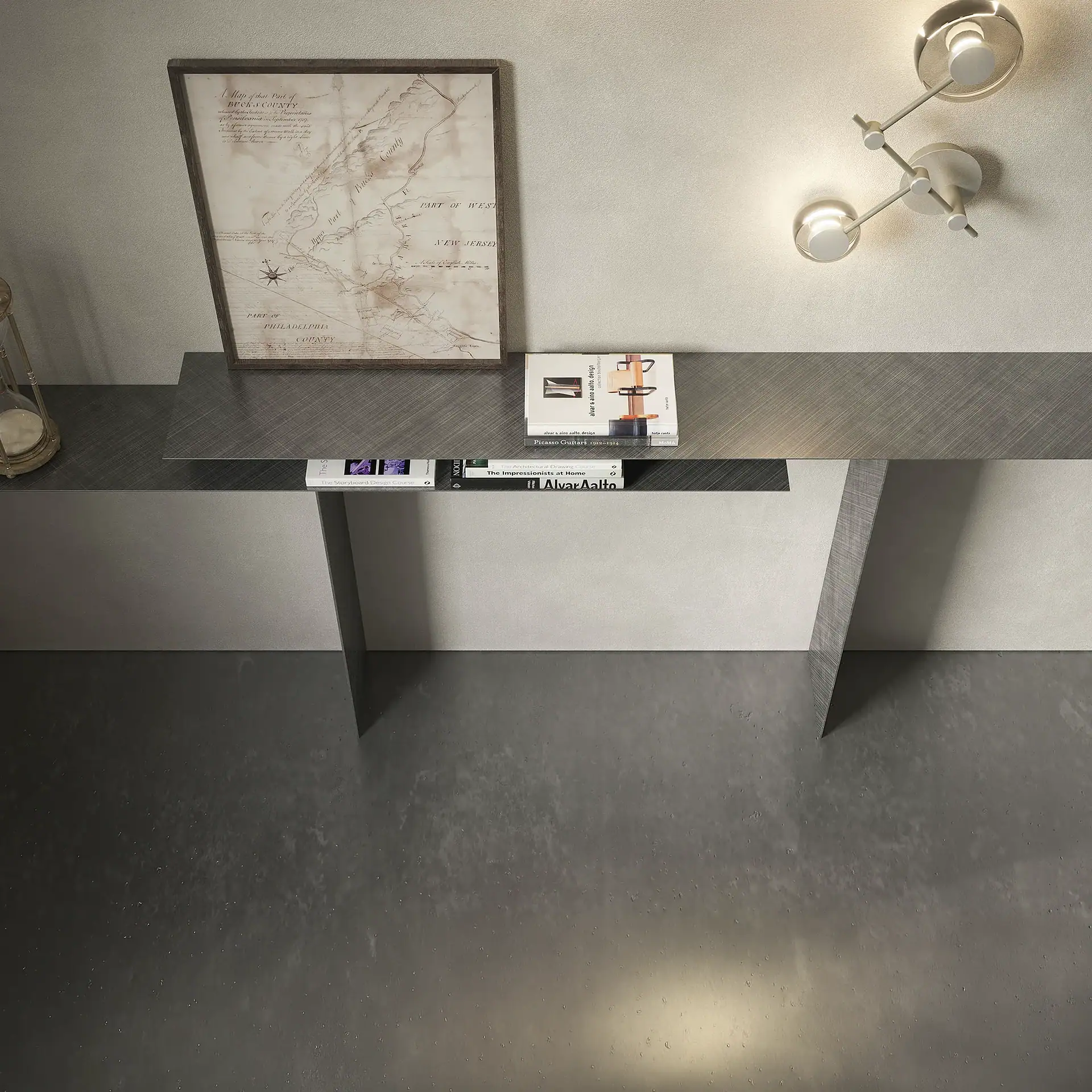 A dark gray console table with a vintage map, books, and an hourglass, illuminated by a modern wall sconce, against a light wall and concrete floor.
