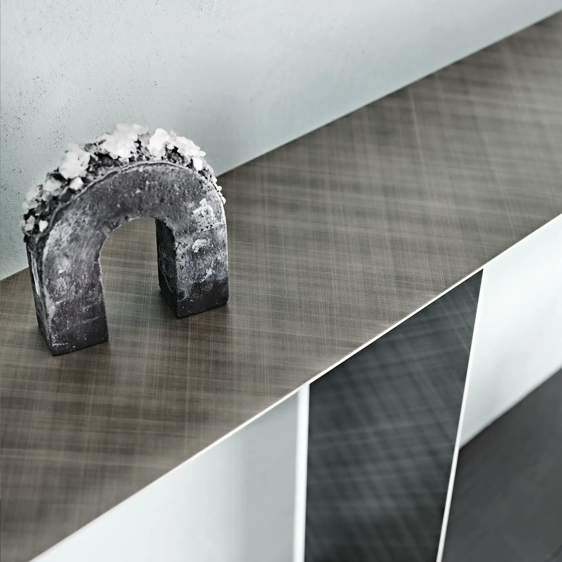 A dark, U-shaped object with white crystals on top, resting on a textured gray shelf against a light wall, with geometric shelving below.
