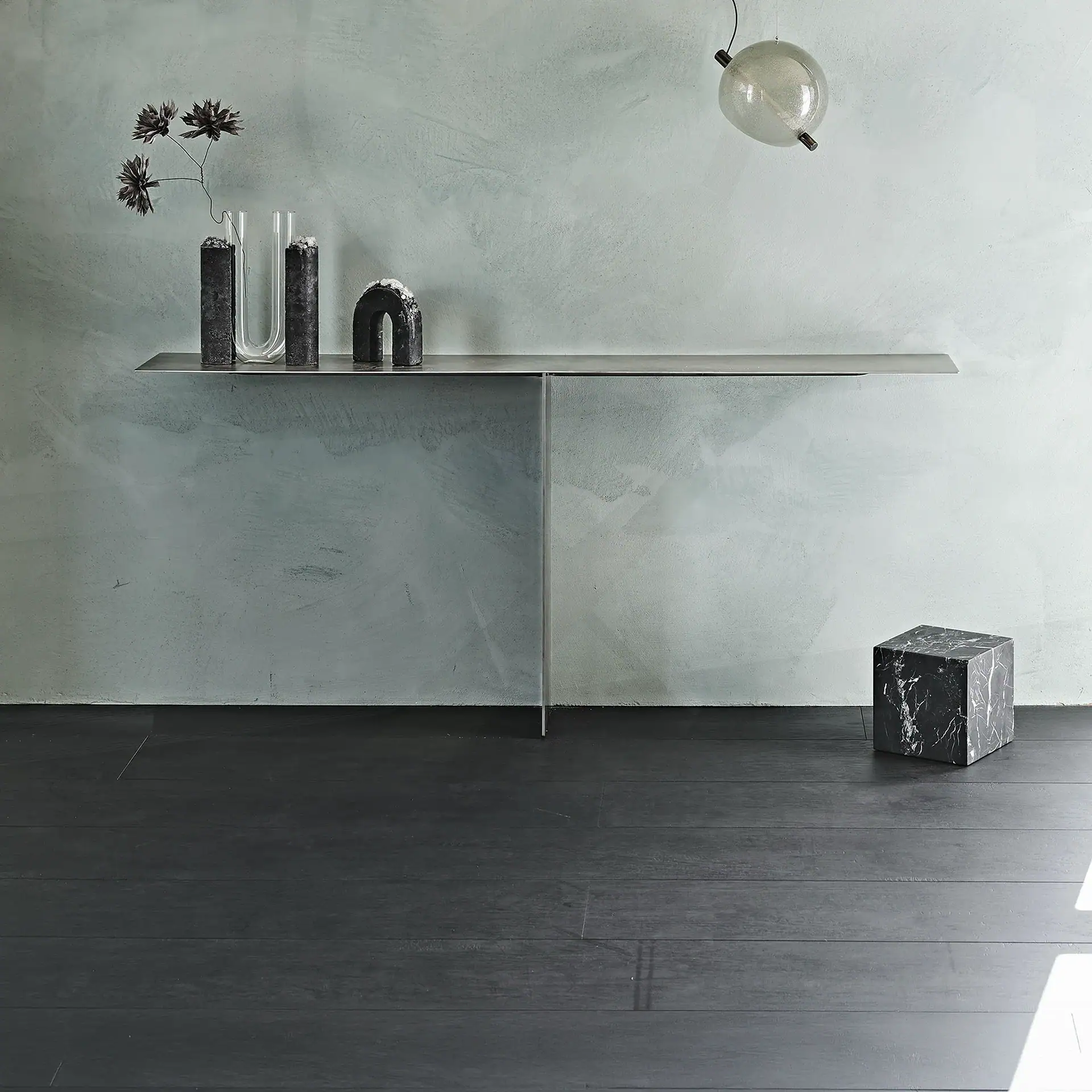 A minimalist console table with decorative objects, including dark blocks, a U-shaped vase with abstract flowers, and an arched sculpture, against a textured light green wall. A dark marble cube sits on a dark wood floor, and a spherical pendant light hangs in the upper right.