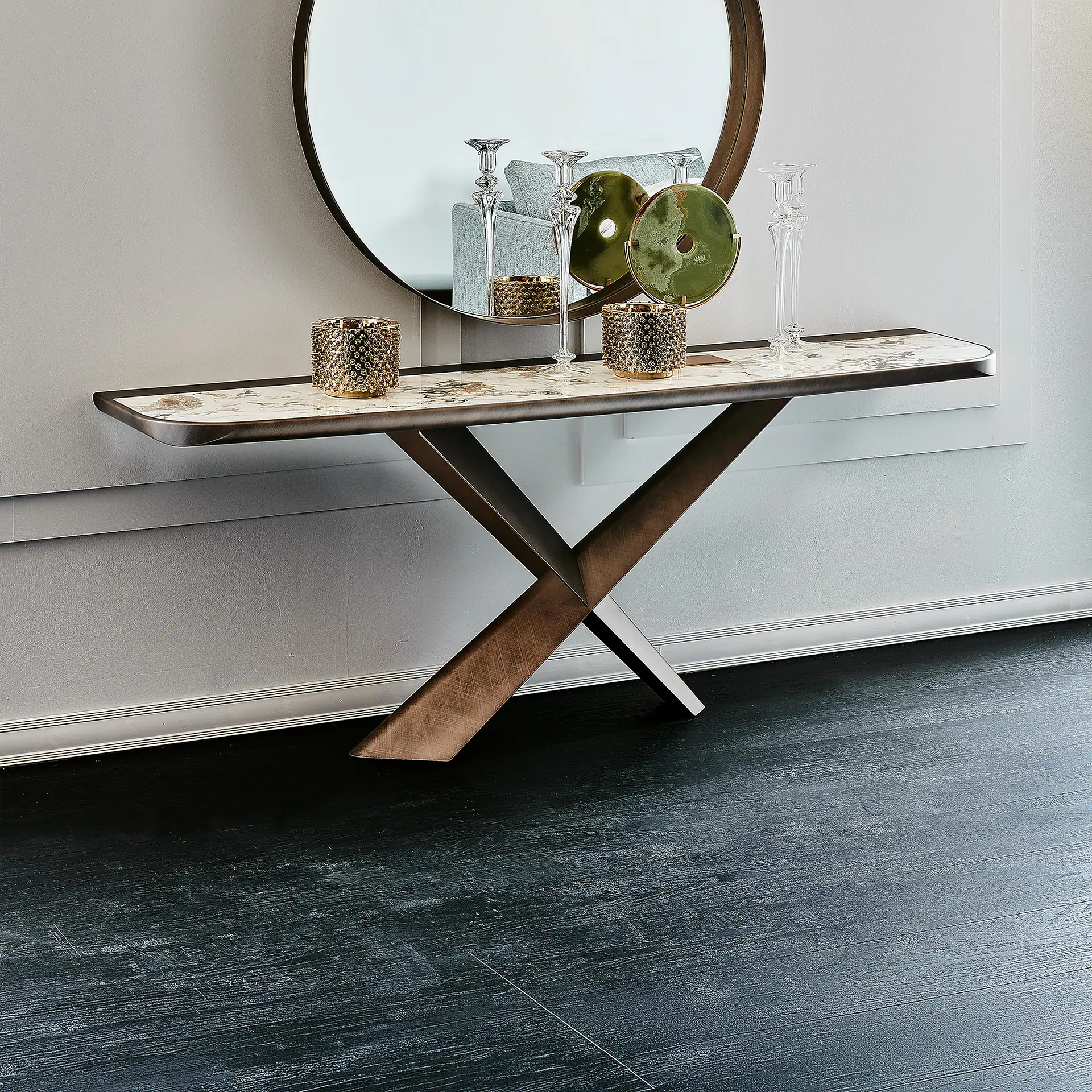 A console table with a marble top and bronze X-shaped legs, adorned with golden candle holders, glass candlesticks, and green circular decor, beneath a large round mirror reflecting a sofa. The floor is dark.