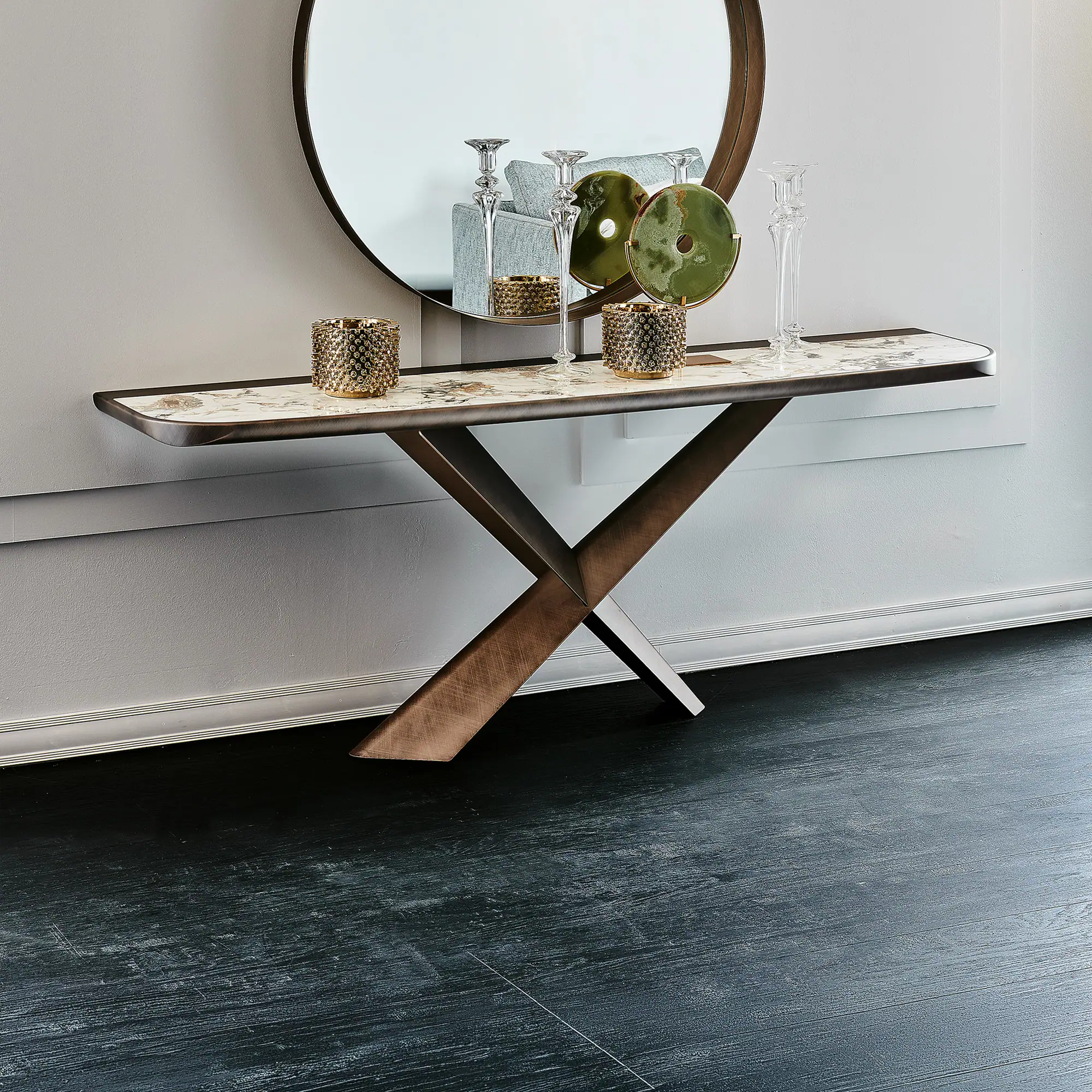 A console table with a marble top and bronze X-shaped legs, adorned with golden candle holders, glass candlesticks, and green circular decor, beneath a large round mirror reflecting a sofa. The floor is dark.