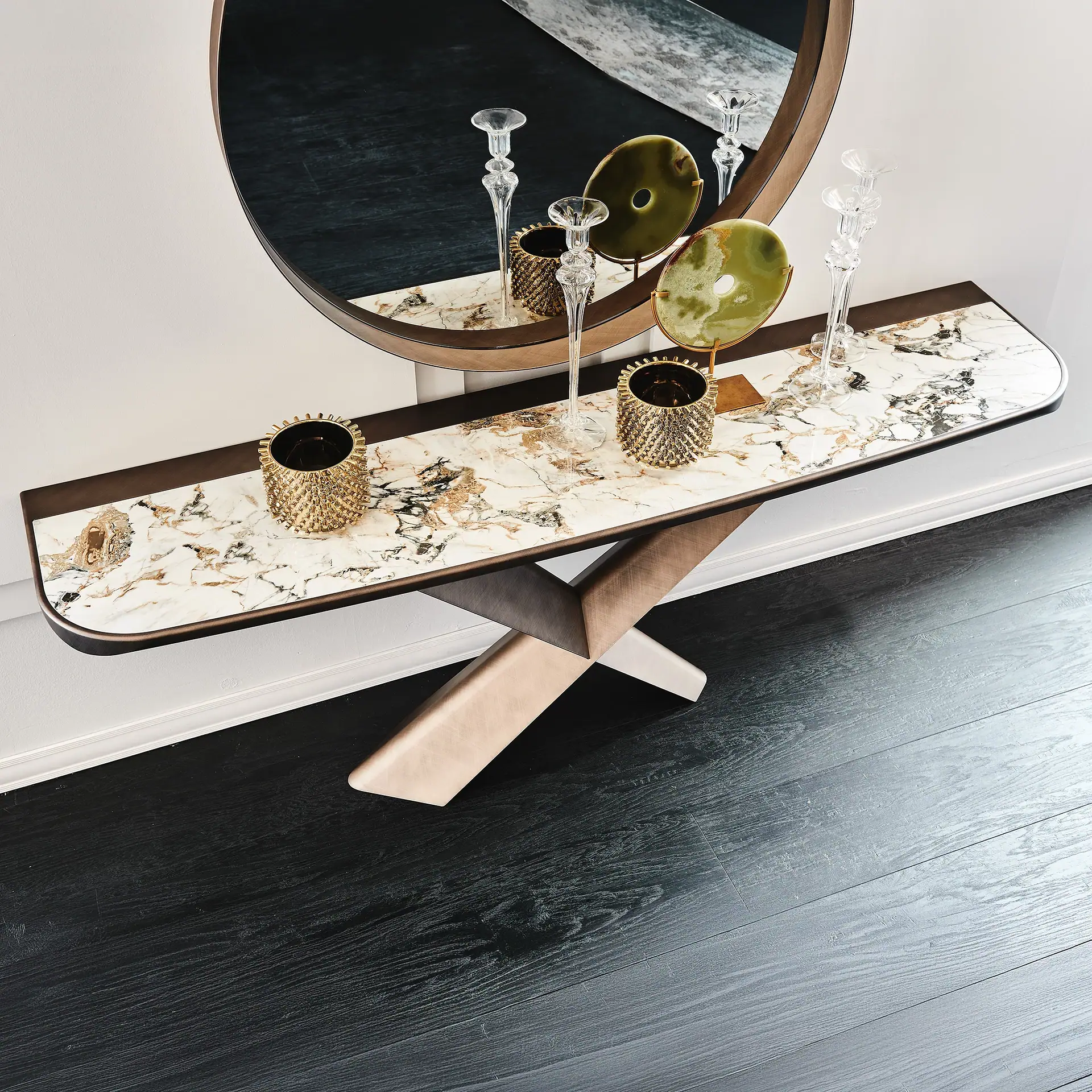 A modern console table with a white marble top and X-shaped legs, paired with a round mirror, decorated with glass candle holders, gold vases, and green circular ornaments, against a white wall and dark wood floor.