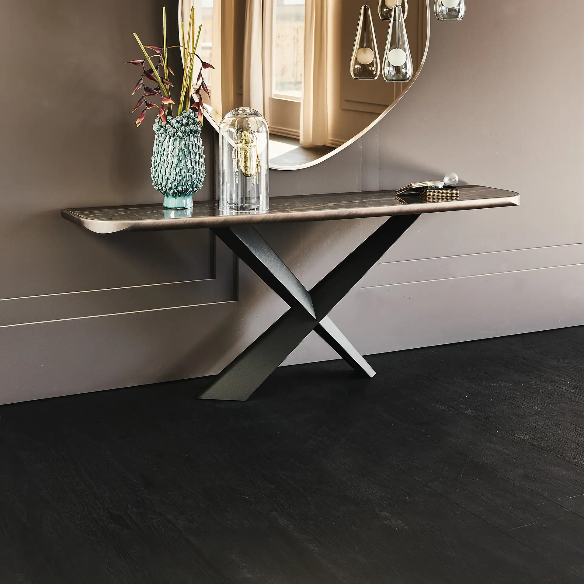 A modern console table with a marble top and X-shaped legs, adorned with a teal vase, a golden beetle under a cloche, and a gold box, set against a dark wall with a large oval mirror reflecting a window.