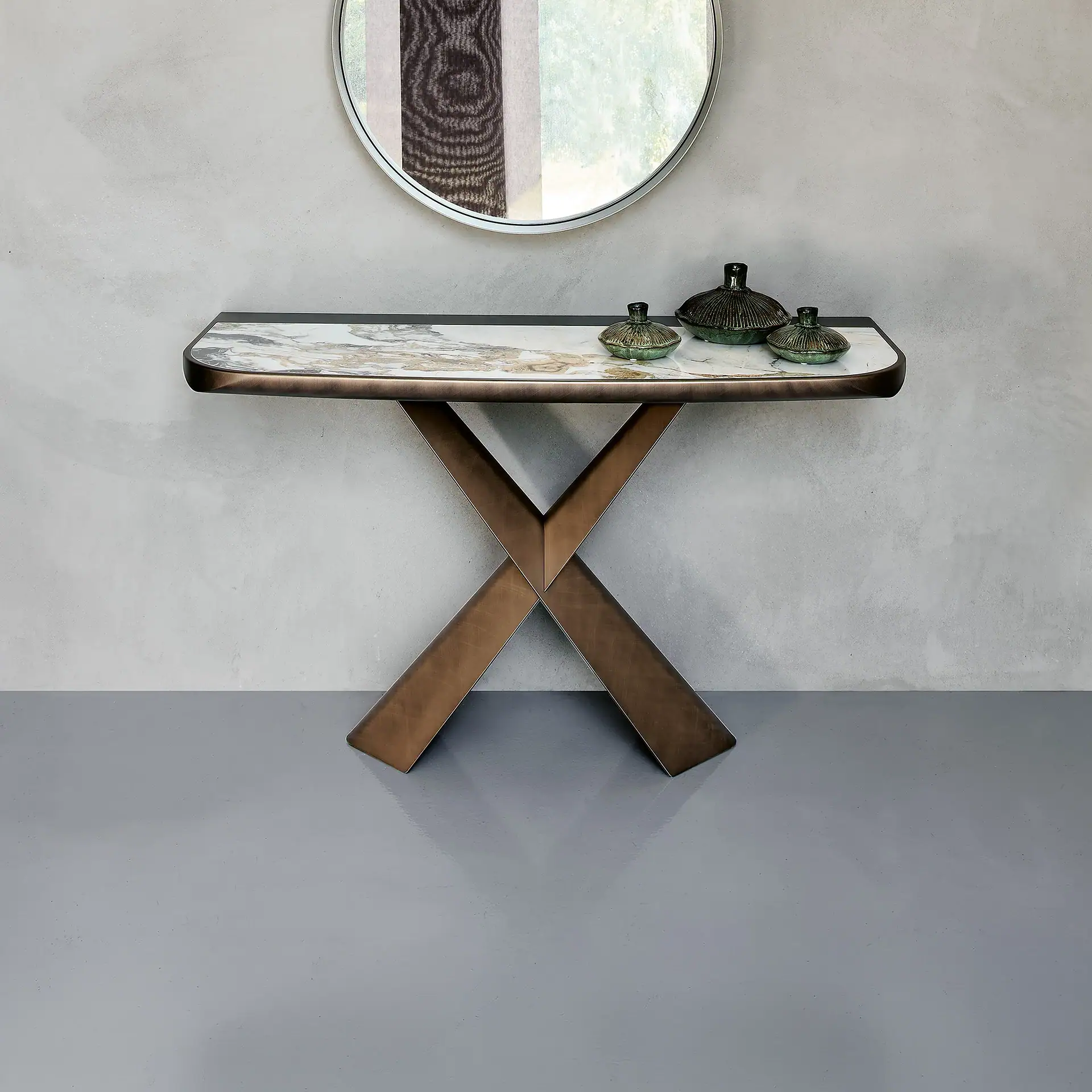 A modern console table with a white marble top and bronze X-shaped legs. Three dark green decorative vessels sit on the table. A circular mirror hangs above, reflecting a window. The wall is light gray, and the floor is glossy gray.