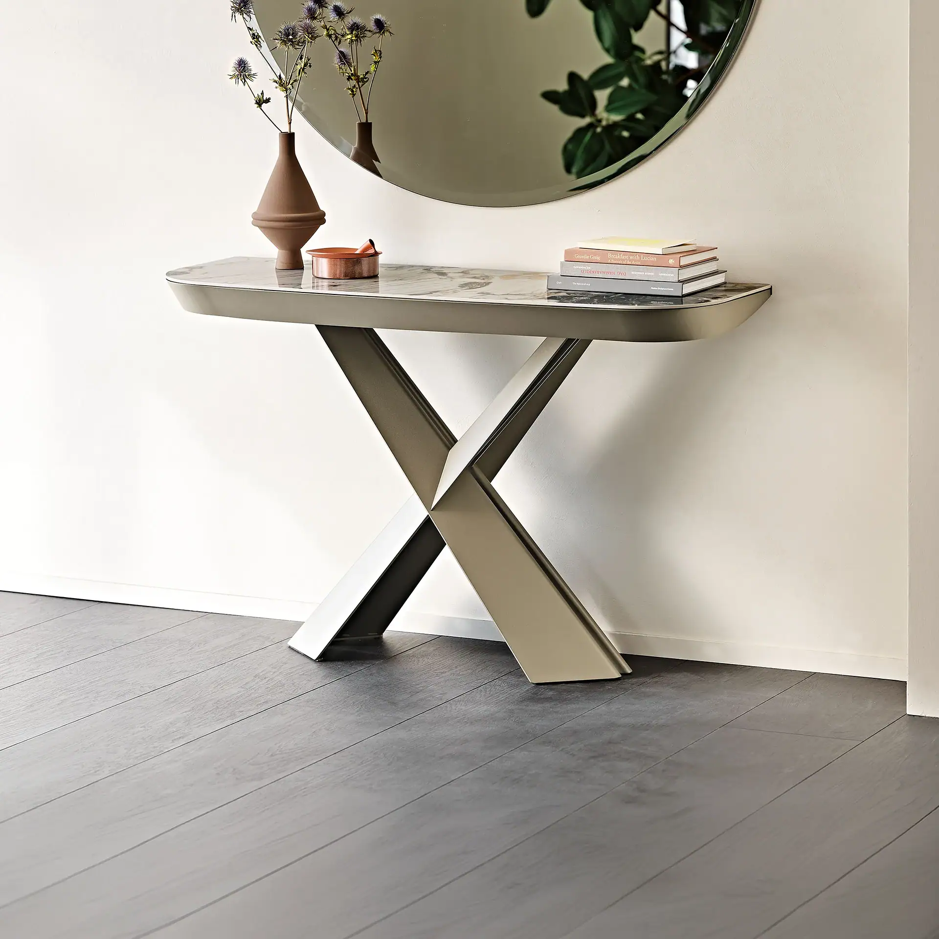 A modern console table with a marble top and X-shaped metallic base, adorned with a vase of thistles, a copper dish, and a stack of books, set against a light wall with a circular mirror reflecting a plant.