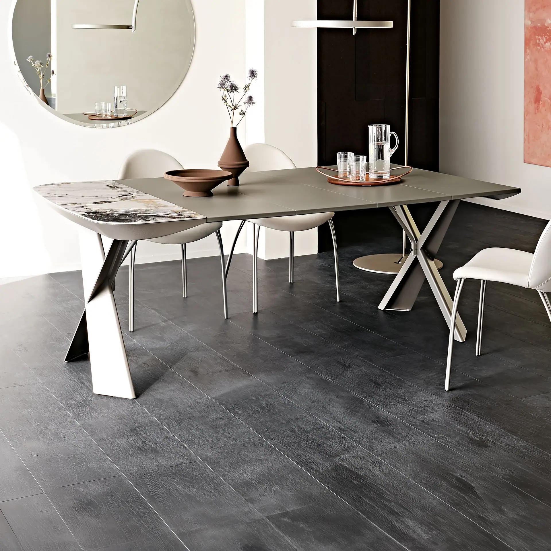 A modern dining room with a gray and marble-patterned table, light chairs, dark wood floor, and a large oval mirror reflecting parts of the room.