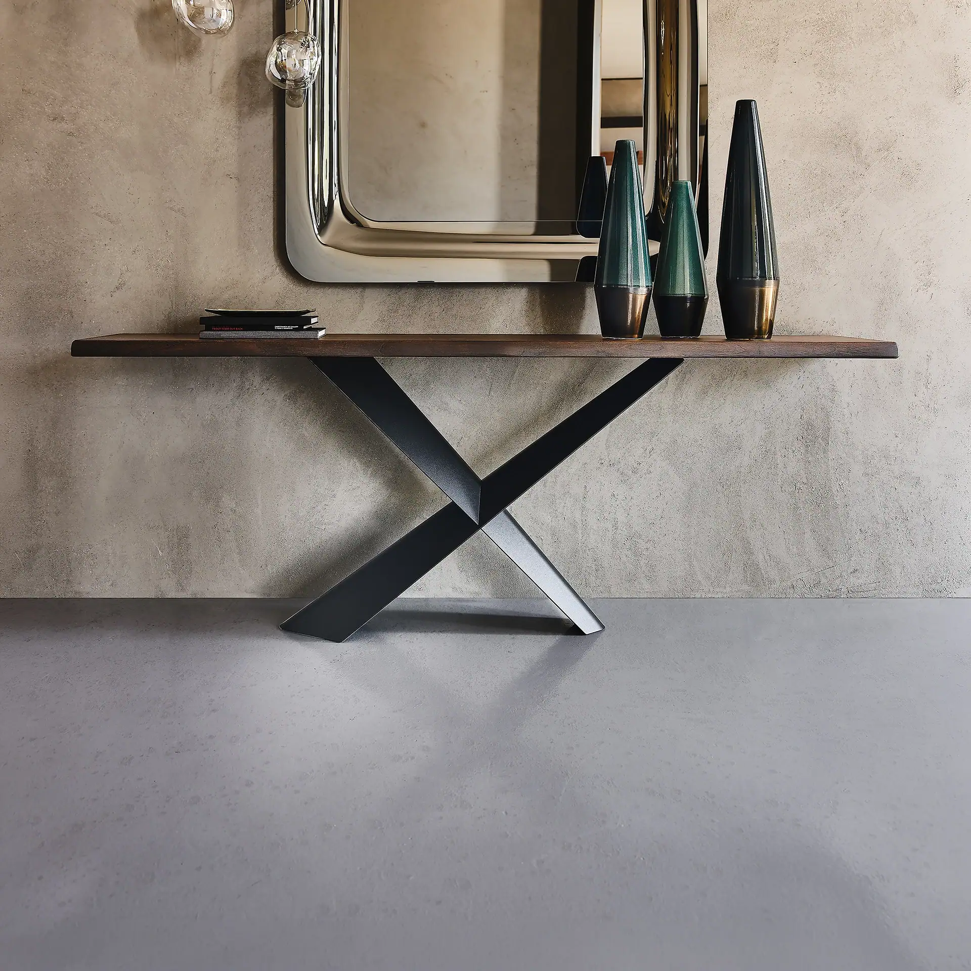 A modern console table with a dark wood top and black X-shaped base against a textured light brown wall. It holds stacked books and three conical teal and bronze vases. A large silver-framed mirror hangs above, reflecting glass pendant lights.