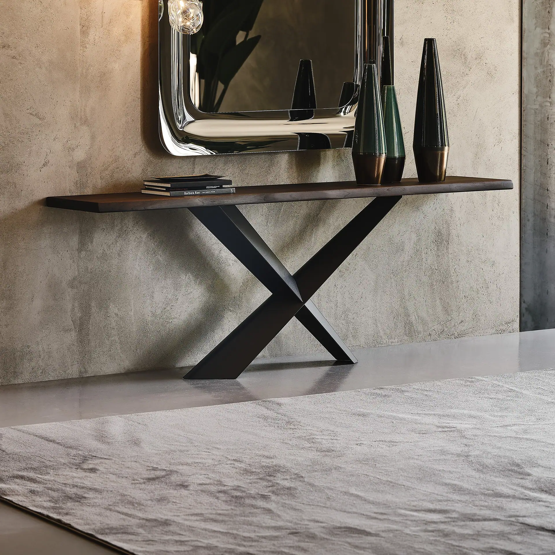 A modern console table with a dark wooden top and X-shaped metal base, adorned with books and three decorative vases, set against a concrete wall with a large chrome mirror.