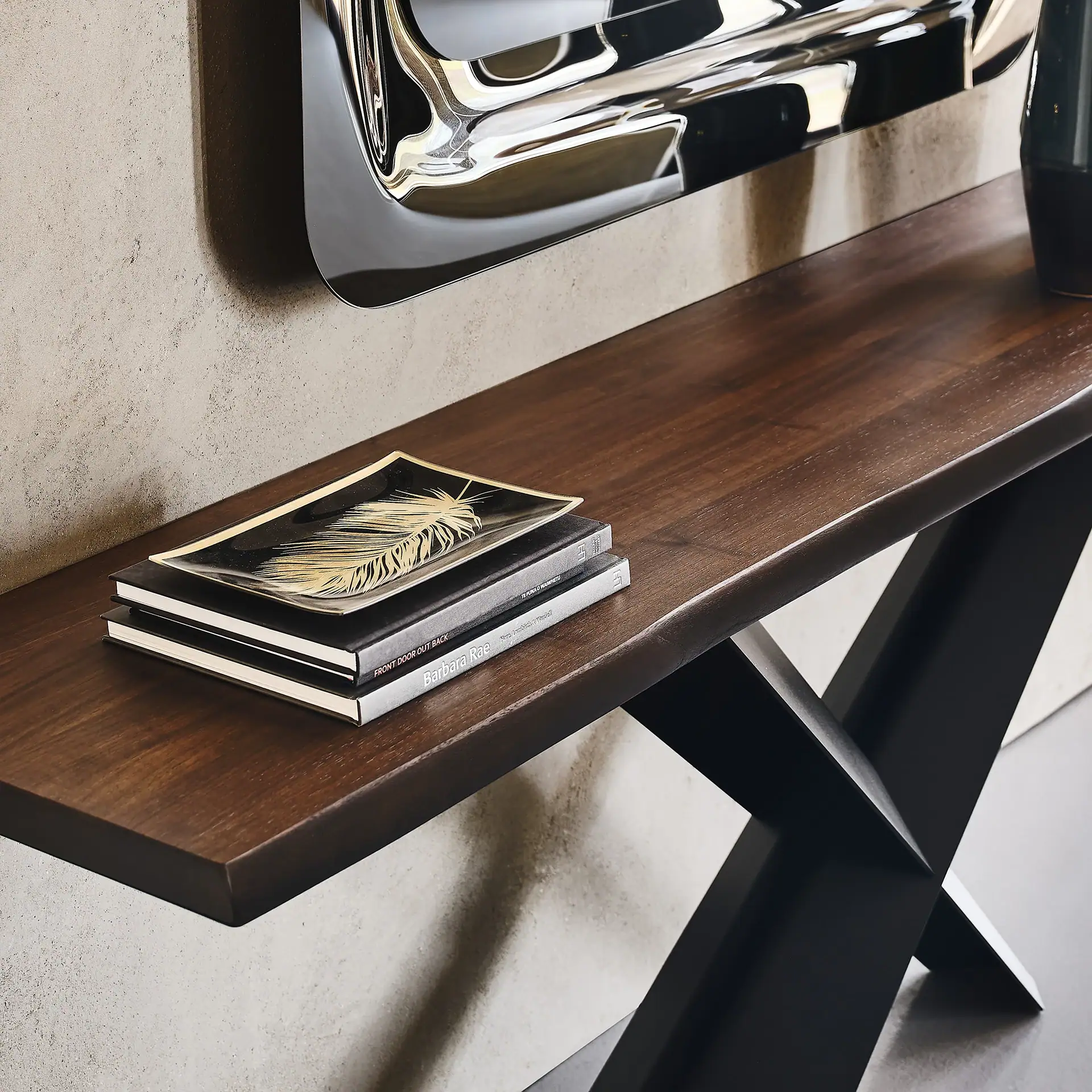 A dark brown wooden console table with black X-shaped legs against a textured gray wall. On the table are two black books topped with a black and gold feather-design tray, and part of a dark vase.