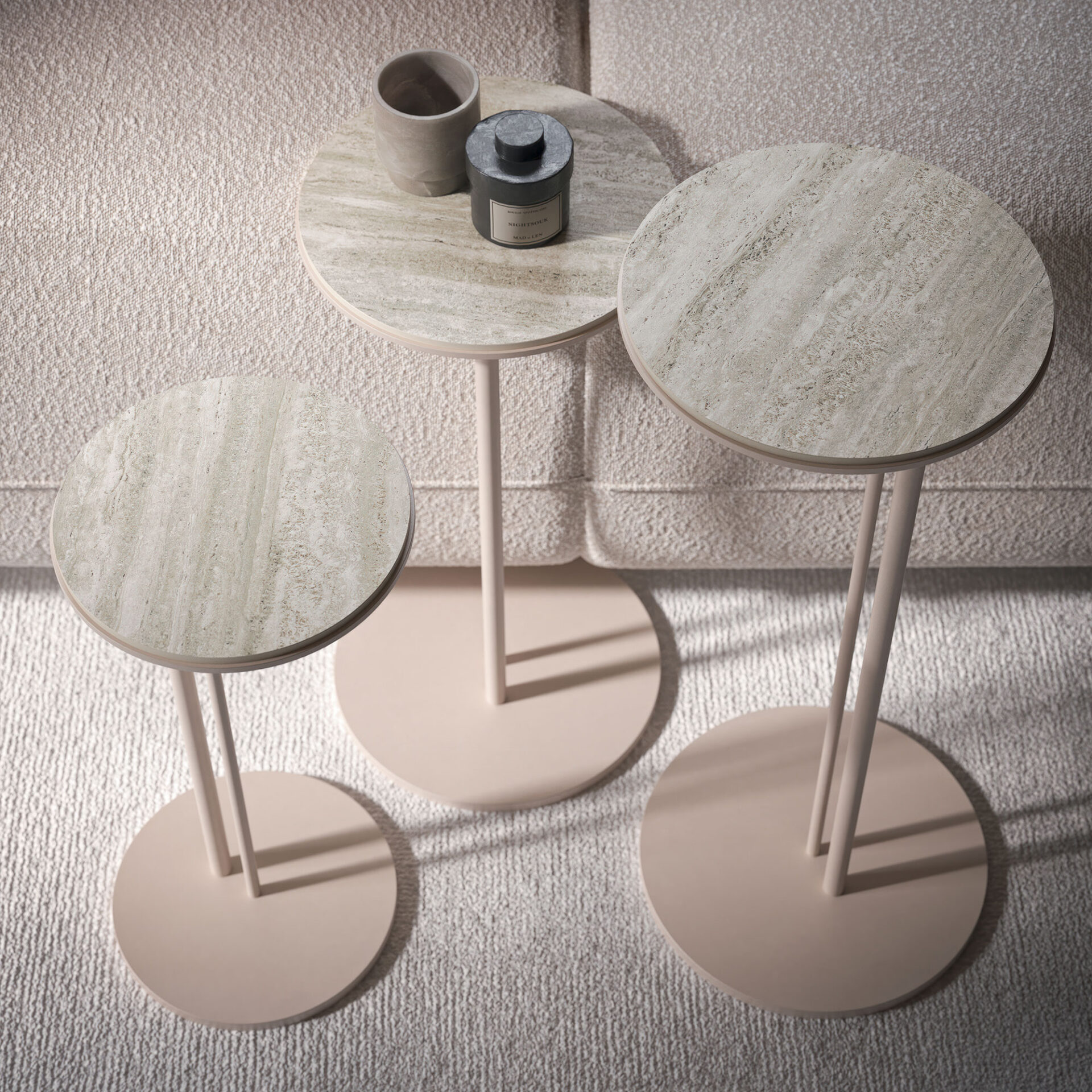 Three round side tables with natural stone tops and light pinkish-beige bases, arranged on a textured rug next to a sofa. One table holds a ceramic cup and a candle.