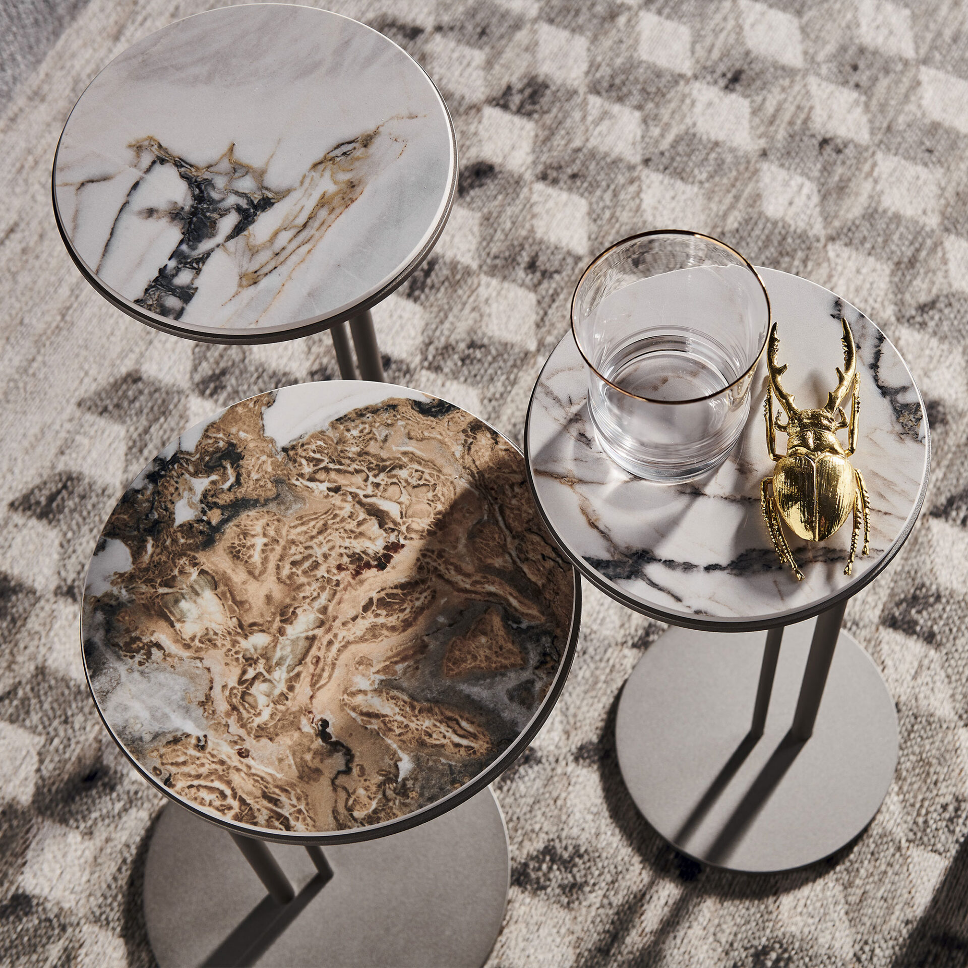 Three round side tables with marble tops on a geometric rug. One table holds a glass with a gold rim and a golden beetle figurine. The marble tops feature white, grey, brown, and gold veining.
