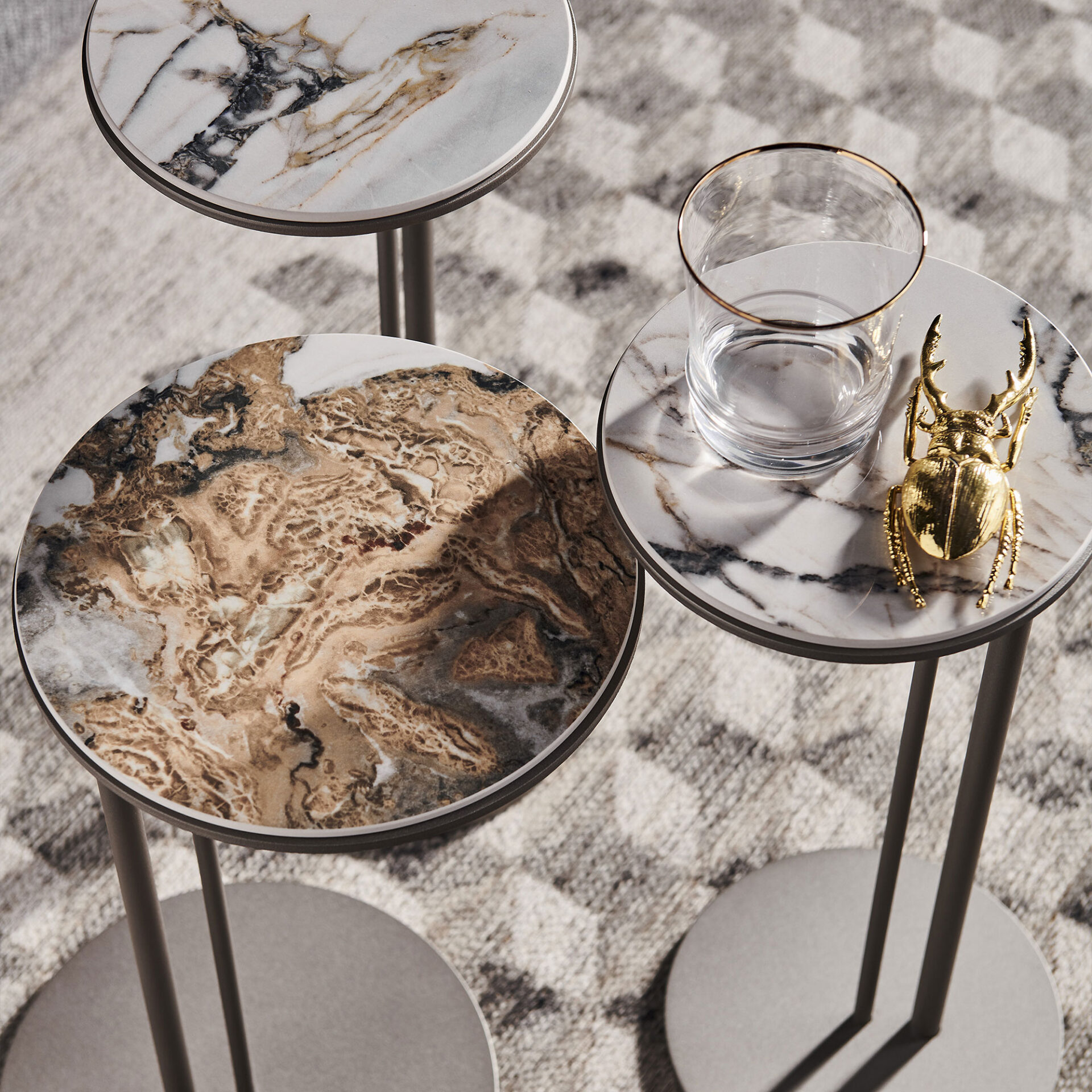 Three round marble-topped side tables on a patterned rug. One table holds a gold-rimmed glass of water and a golden decorative beetle.