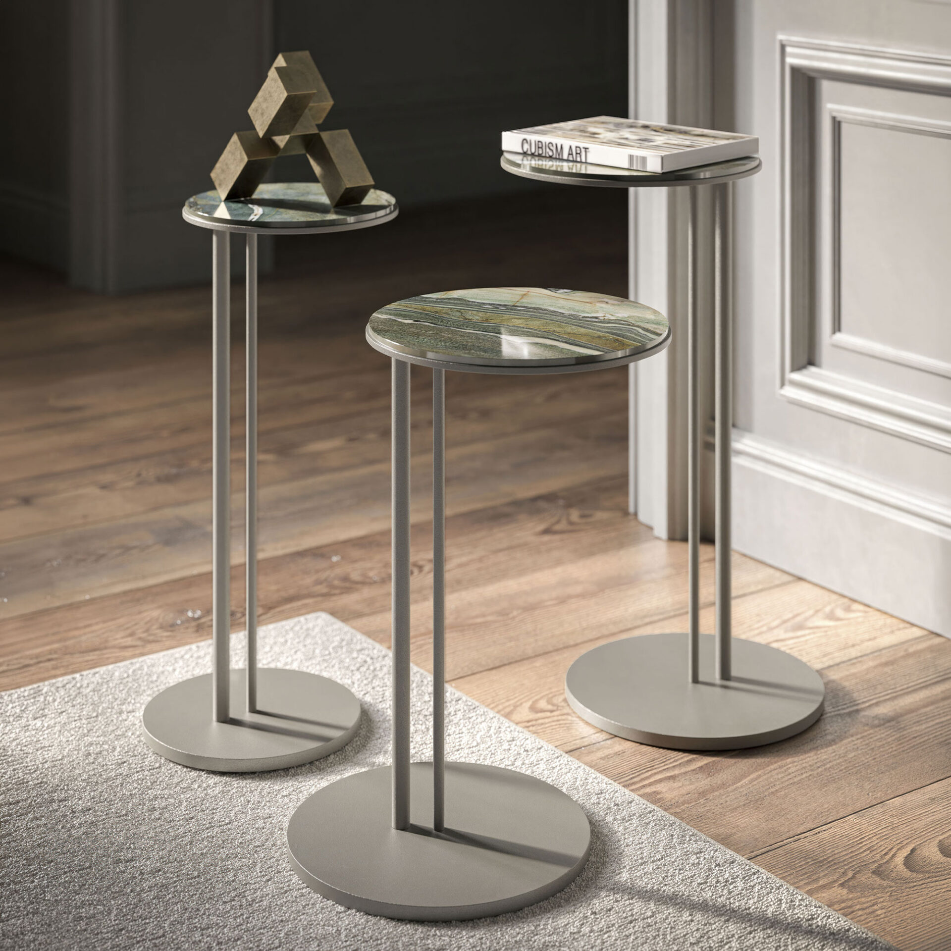 Three modern side tables with circular tops and bases, in a room with a rug and wooden floor. The tables have marble and plain grey tops, one holding a brass geometric sculpture, another a book titled 'CUBISM ART'.