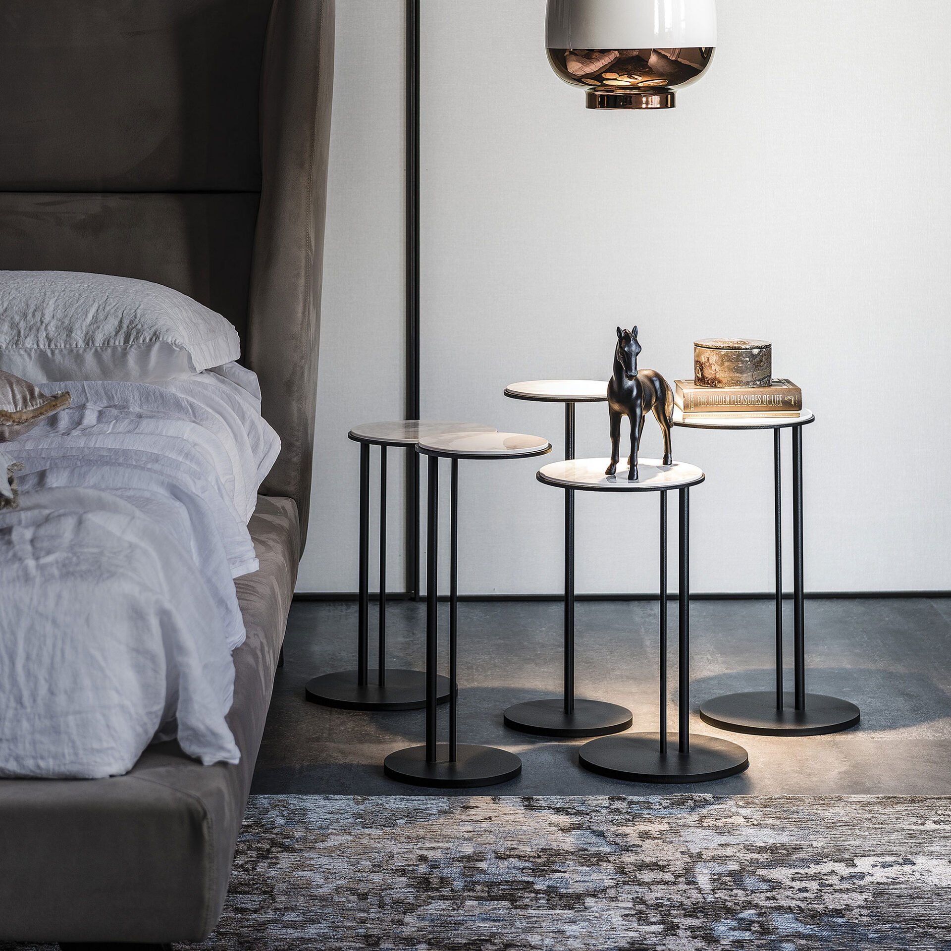 A modern bedroom with a dark upholstered bed, white linen, and three marble-topped side tables holding a horse figurine, a decorative box, and a book. A pendant light hangs above.